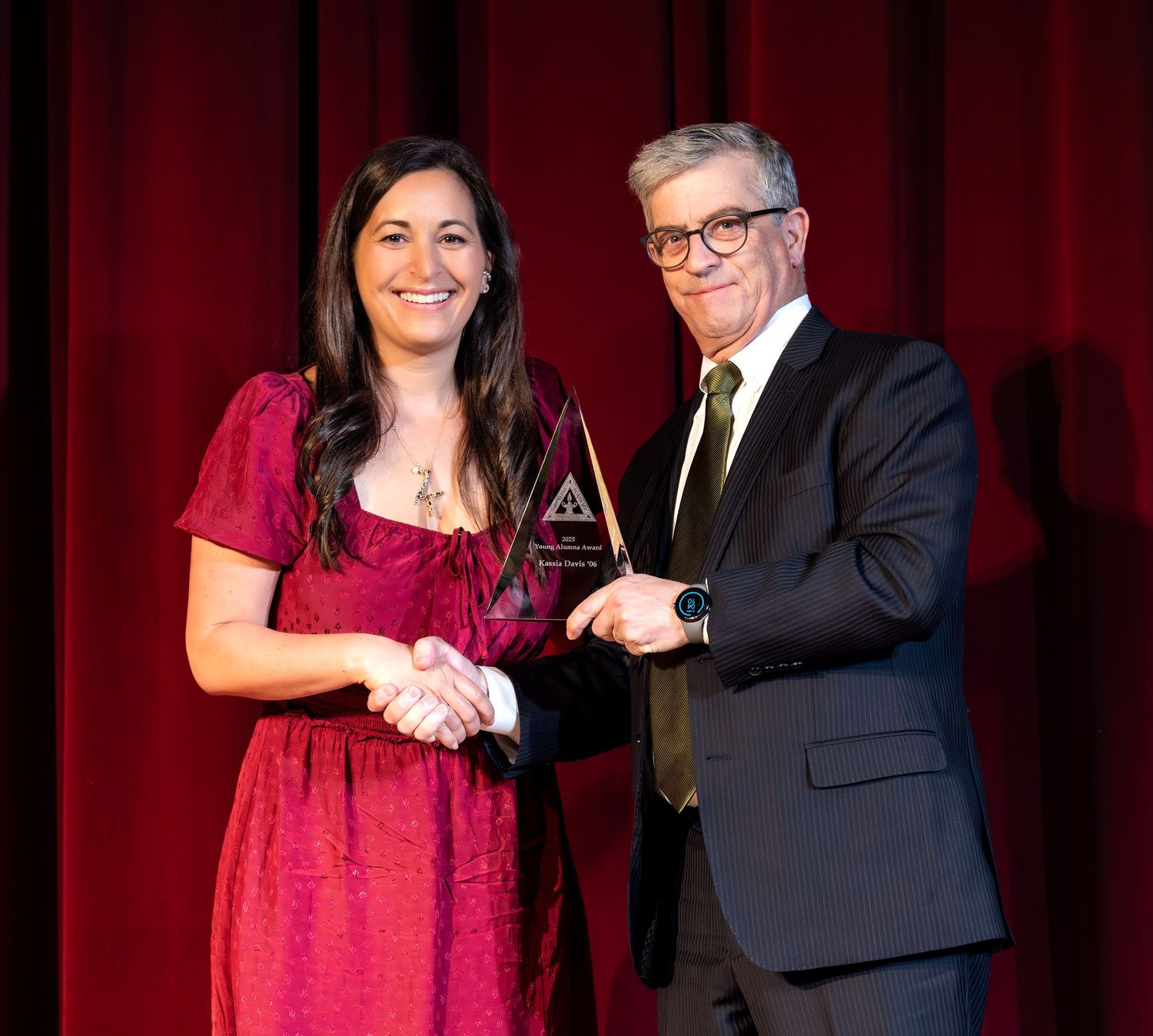 A person in a red dress receives an award trophy from someone in a dark suit on a stage with red curtains.