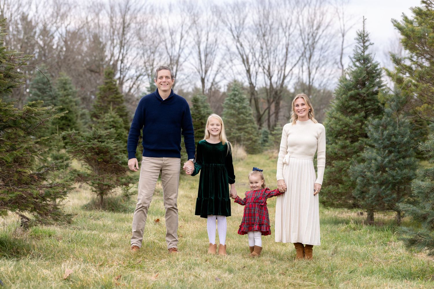 A family stands together outdoors among evergreen trees at a Christmas tree farm on a cloudy winter day.