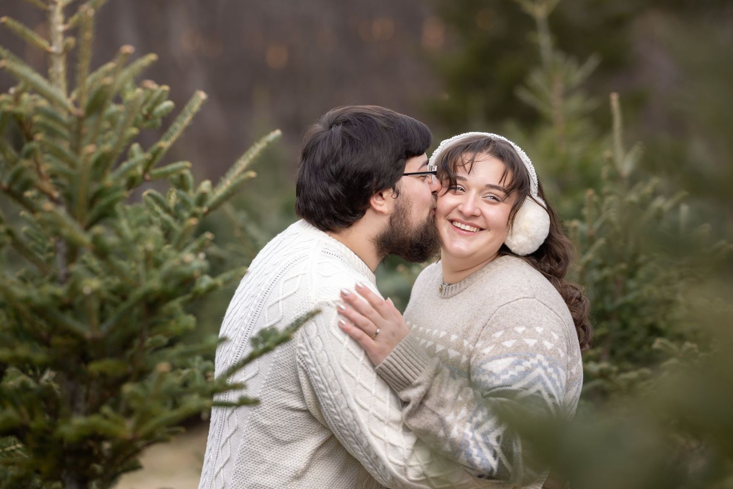 A couple in white sweaters sharing an intimate moment among evergreen trees at a Christmas tree farm.