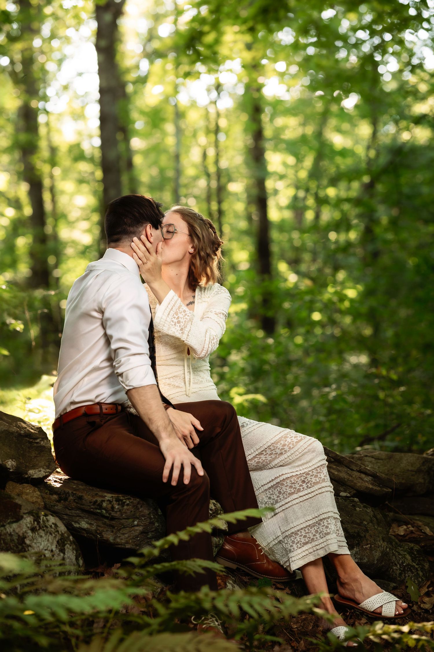 A romantic couple sits together on a fallen tree trunk in a sunlit forest setting.