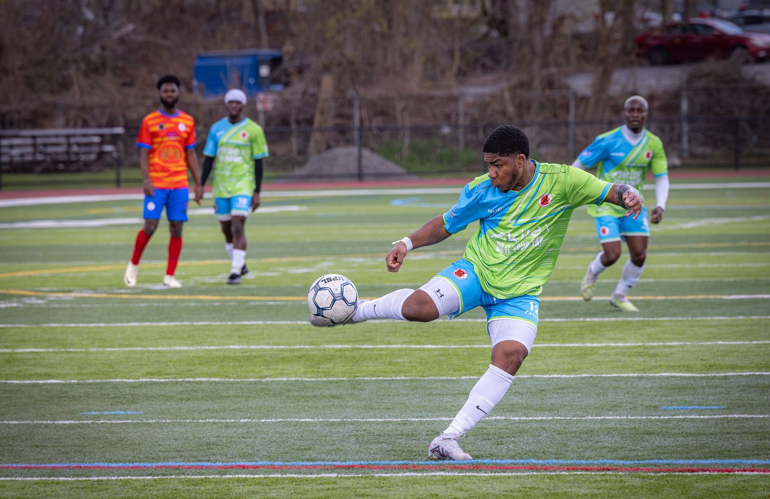 Soccer players in colorful jerseys practicing shots and drills on an outdoor turf field.