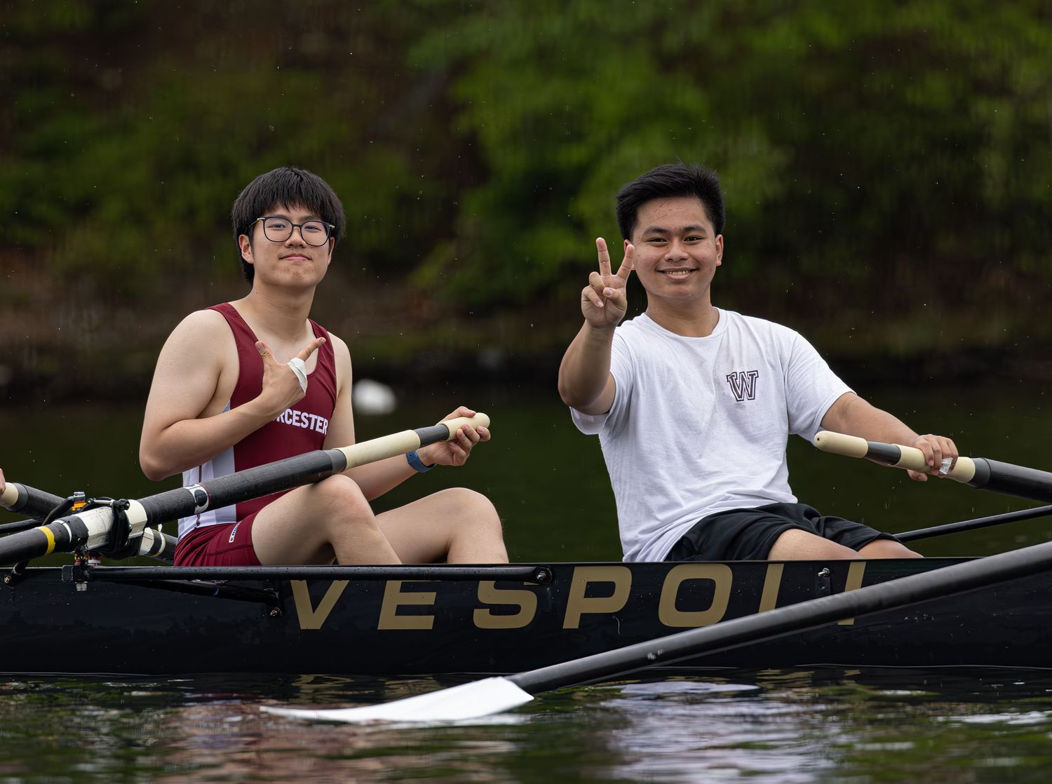 Two rowers in a Vespo boat paddle together on calm waters with trees in the background.