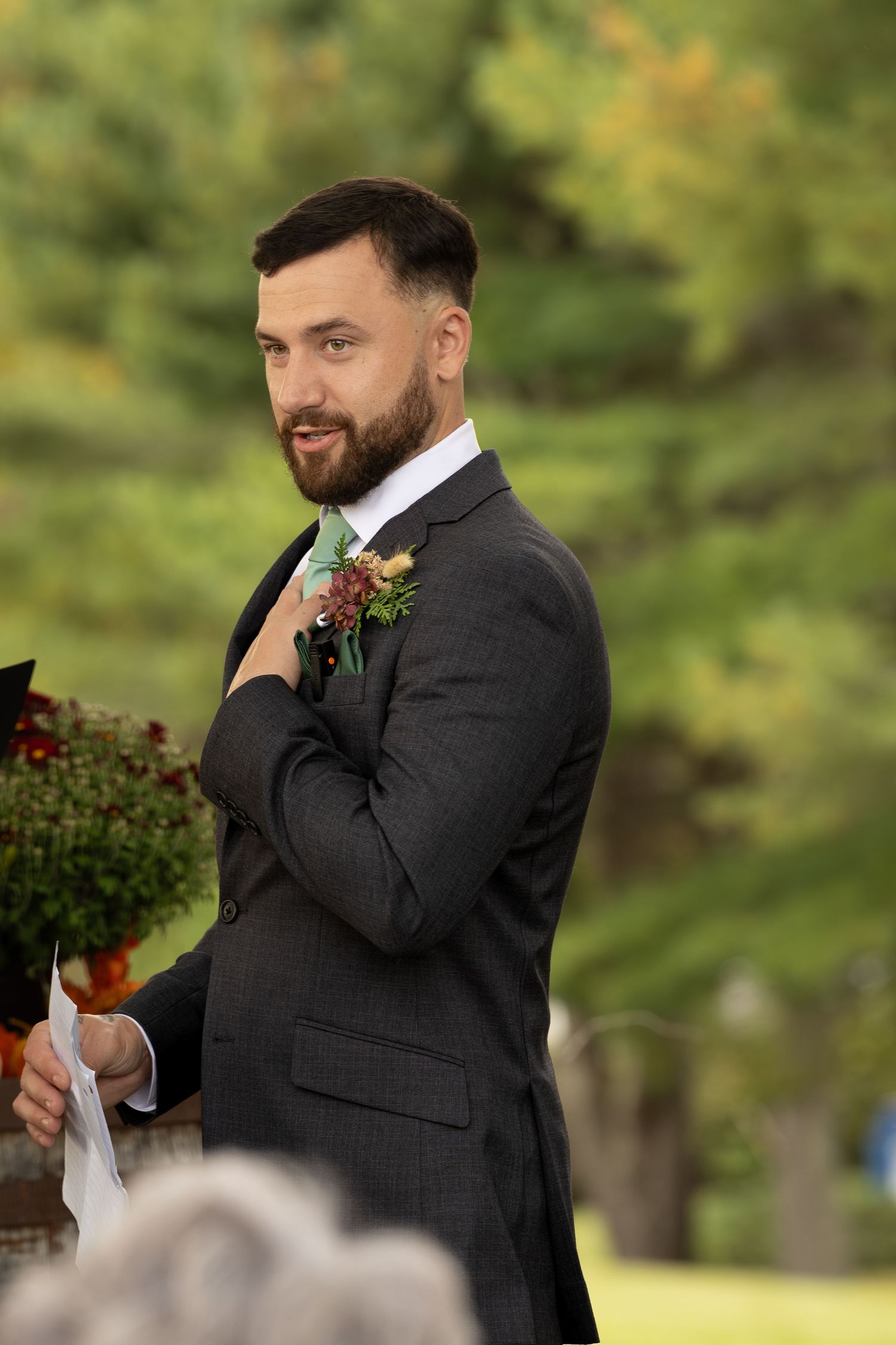 A groom in a charcoal suit adjusts his tie while wearing a burgundy boutonniere during an outdoor autumn wedding ceremony.