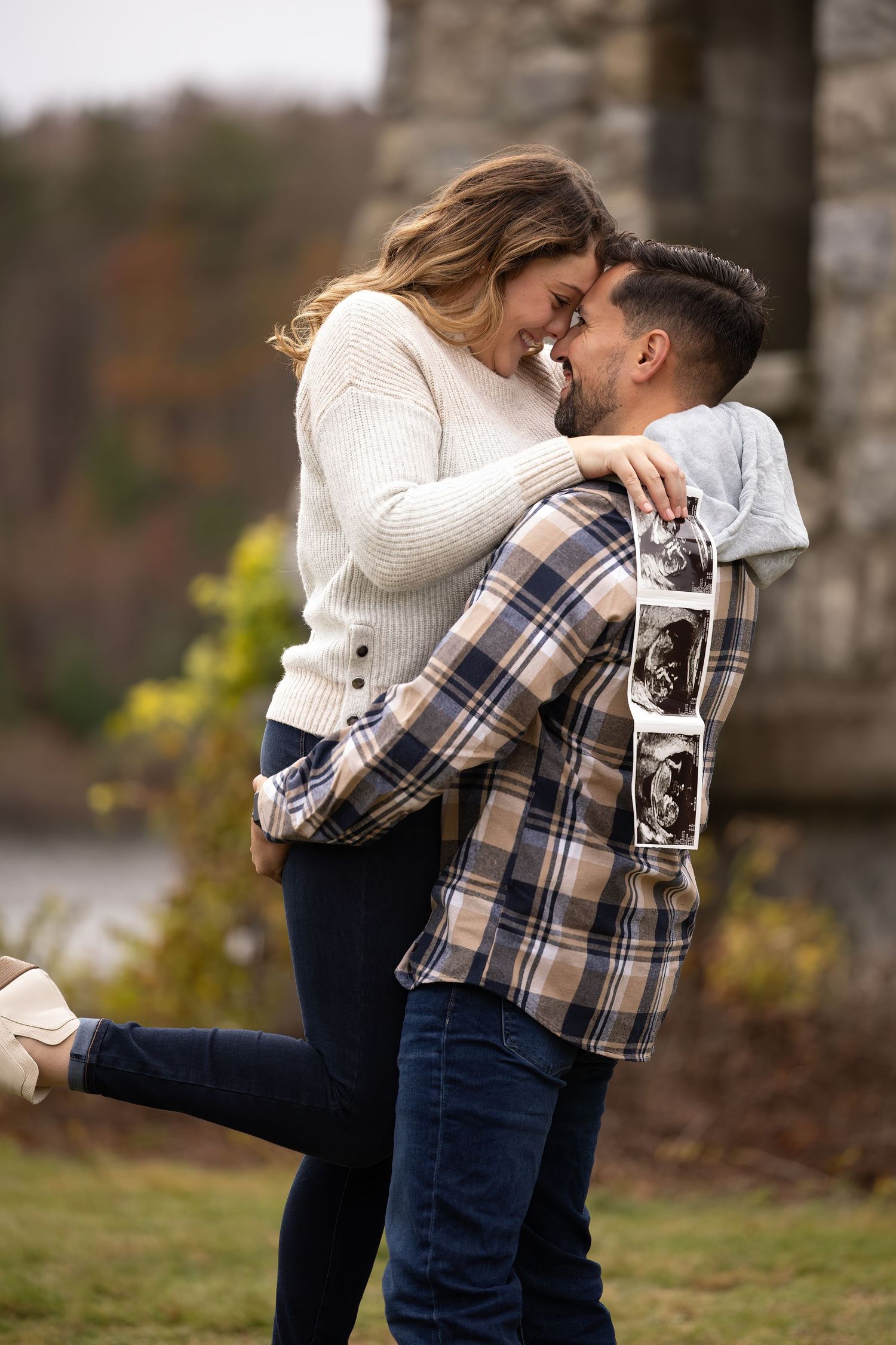 A couple in coordinated white sweater and plaid shirt embrace while holding a vintage camera outdoors.