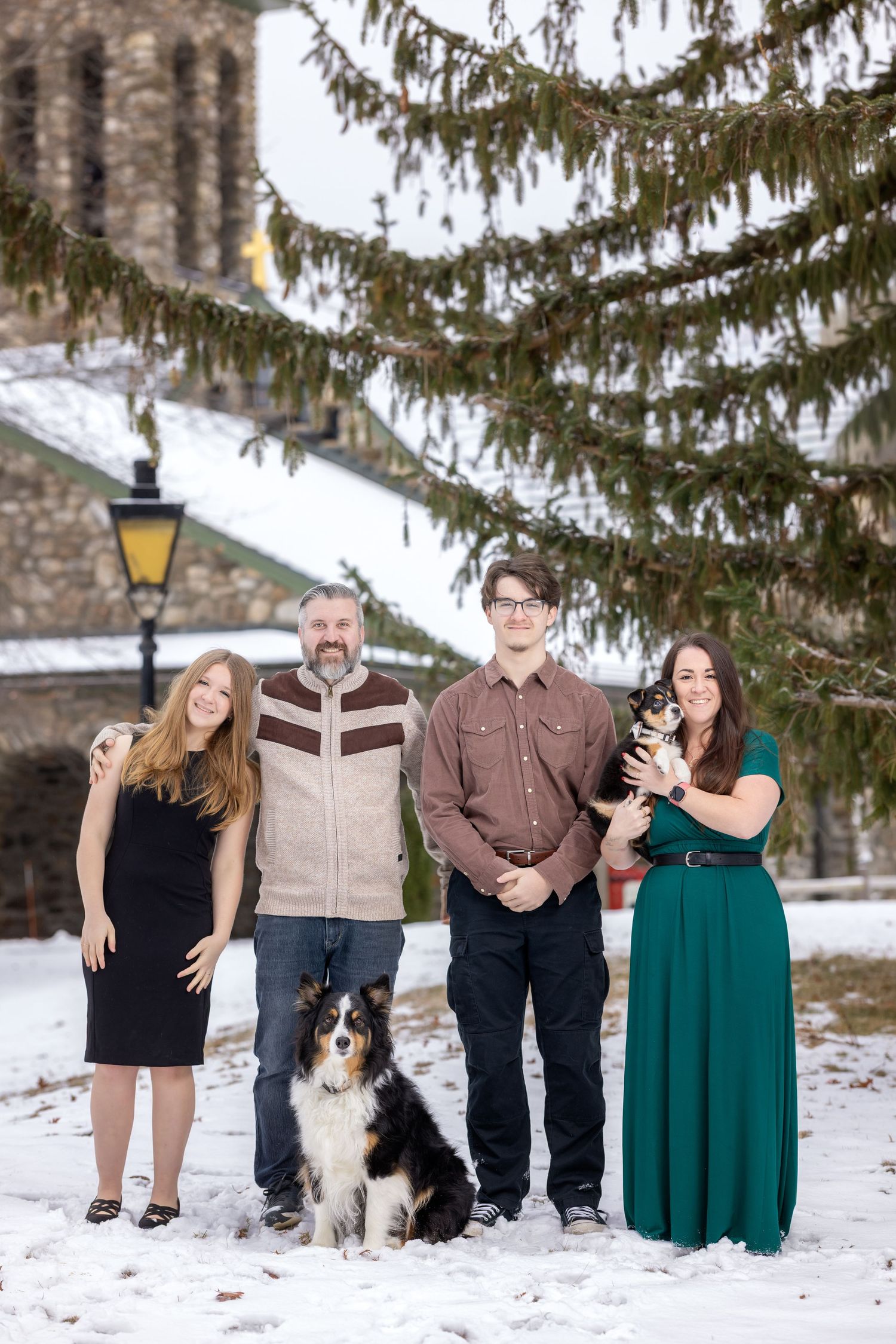 A family poses with their black and white Border Collie dog in a snowy winter setting under evergreen trees.