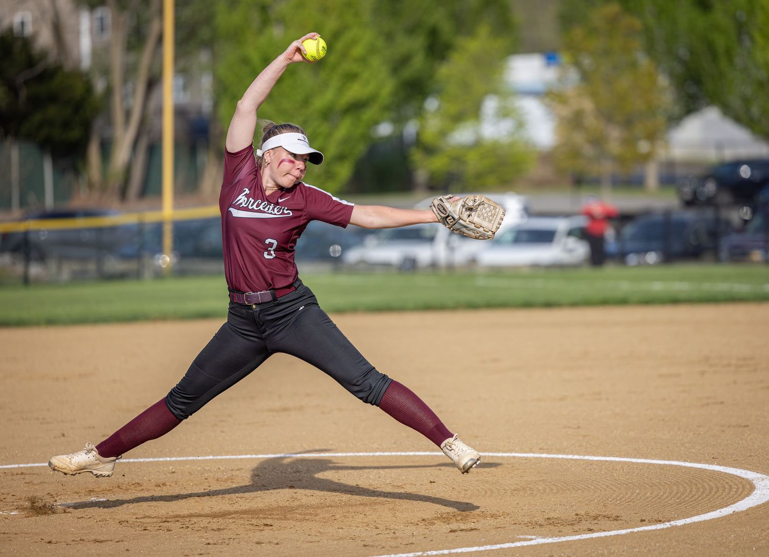 A softball pitcher in maroon uniform winds up to throw during a game on a sunny day at the baseball diamond.