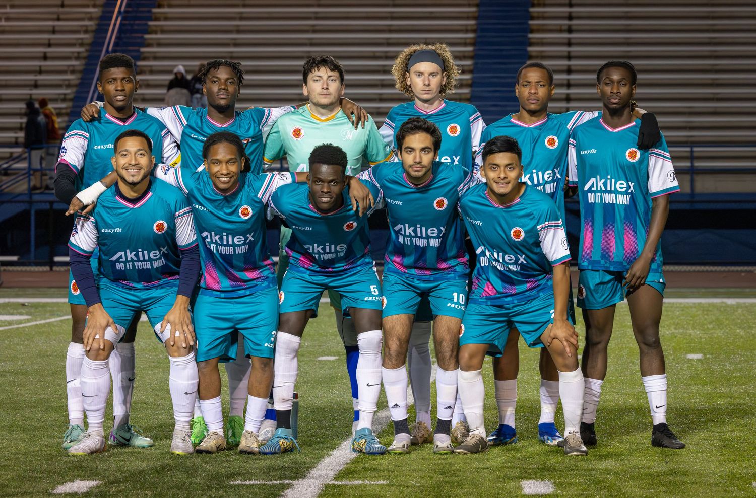 A soccer team poses together for a group photo wearing turquoise blue uniforms on a sports field at night.