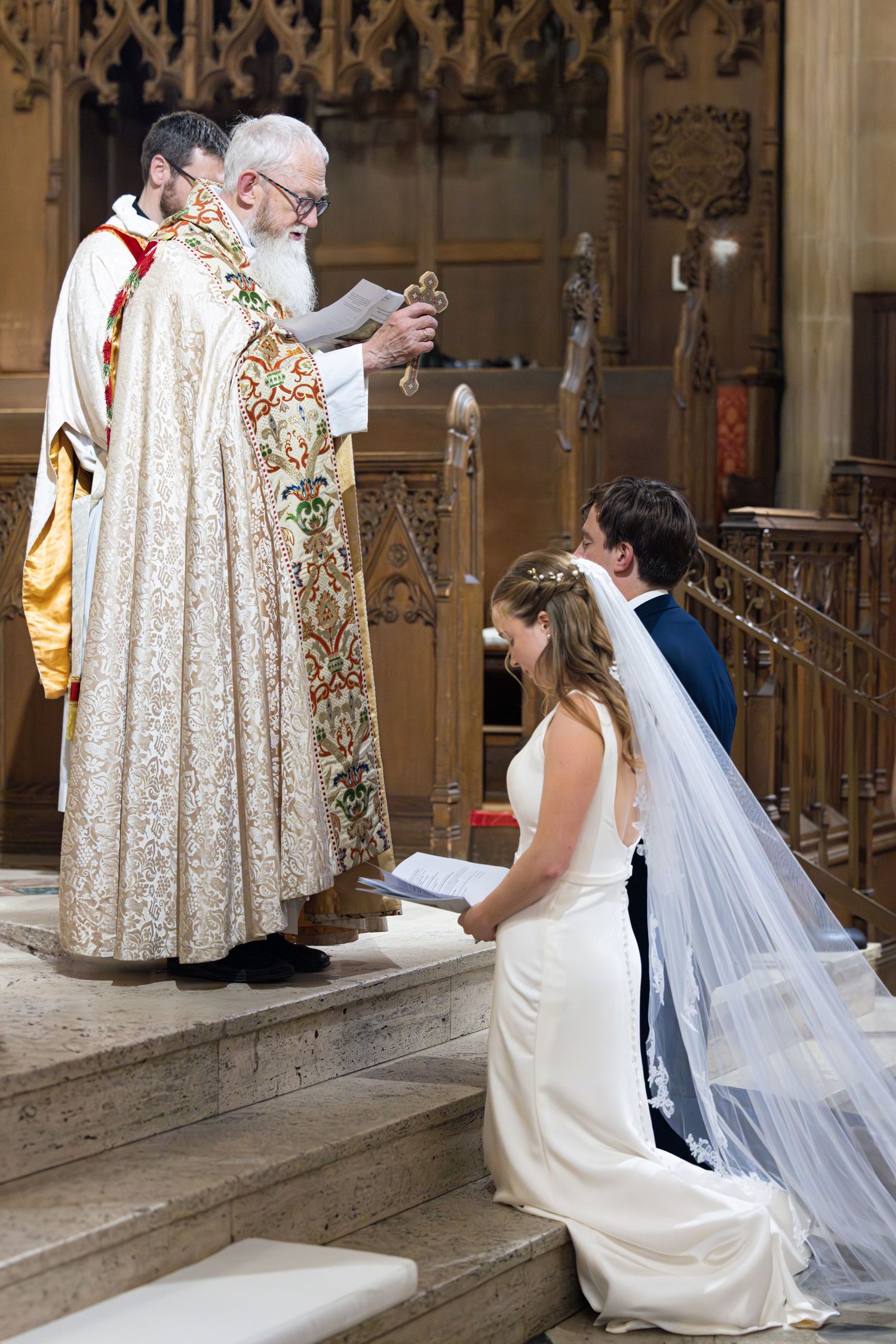 A traditional wedding ceremony takes place on church steps with a priest in ornate robes blessing a kneeling couple.