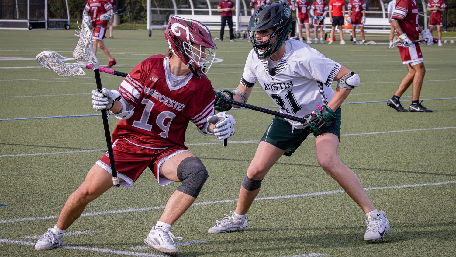 Athletes in red and white uniforms compete in a lacrosse match on an outdoor turf field.