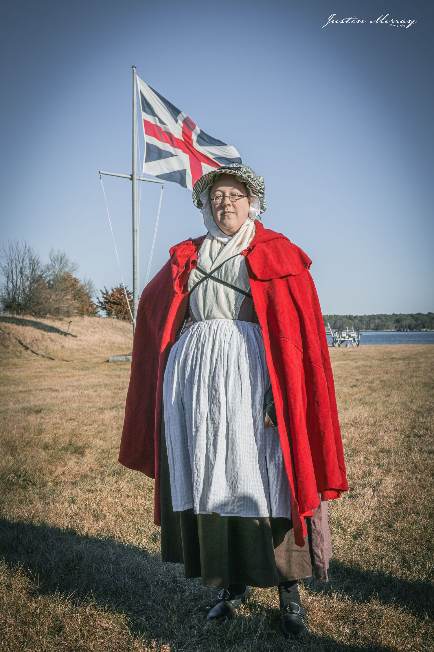 Historical reenactor in traditional colonial dress with red cape stands against coastal landscape with flag pole in background.