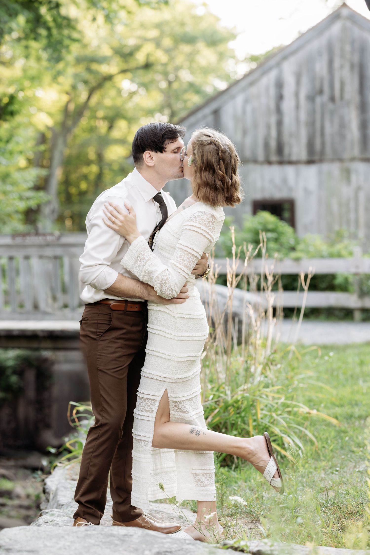 A romantic couple embraces in front of a rustic barn with lush greenery in the background.