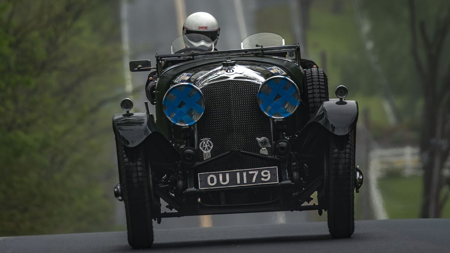 Vintage race car driving on a winding road through lush green trees during a classic car rally event.