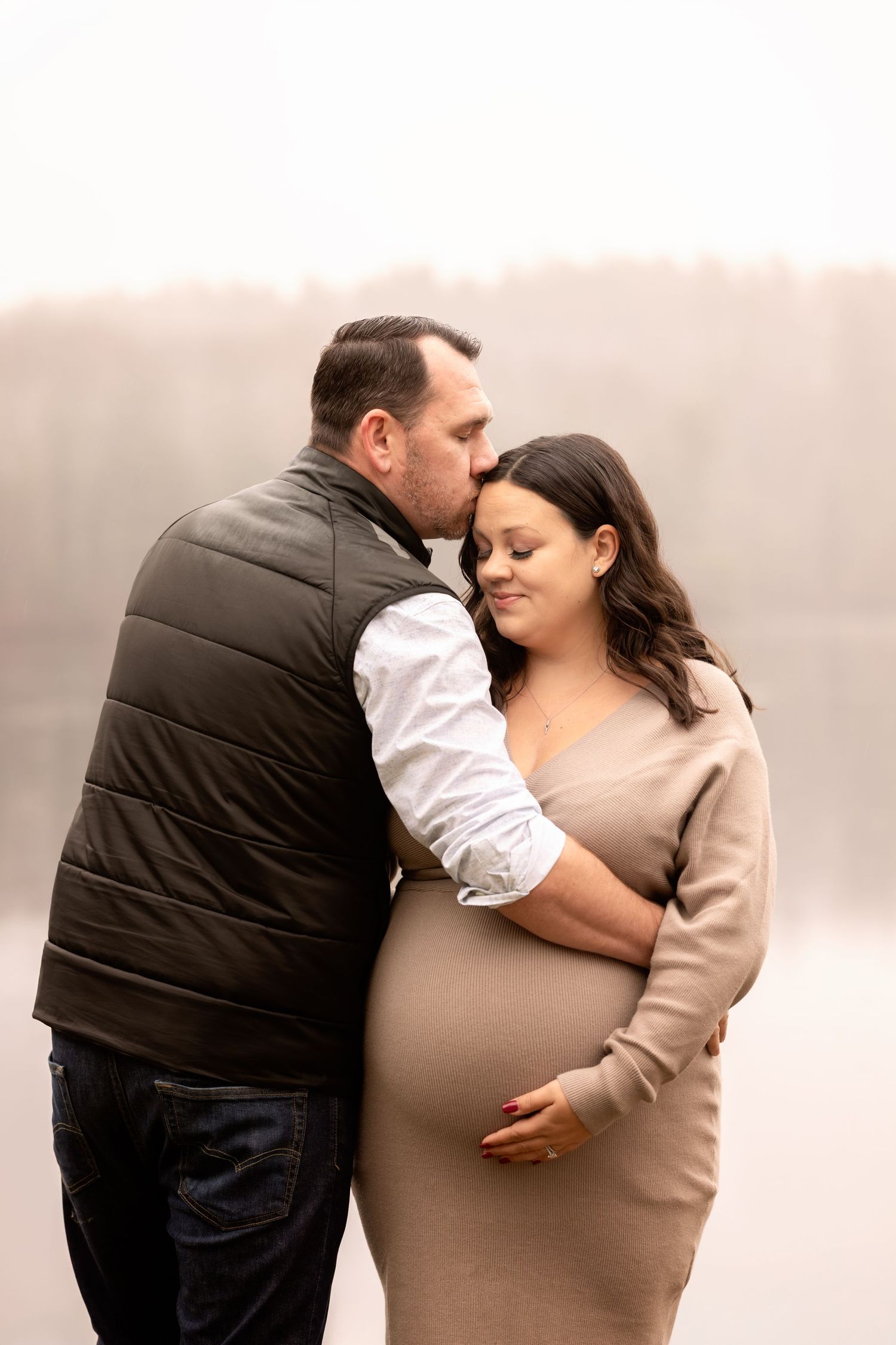 A couple shares an intimate moment during a maternity photo shoot wearing neutral tones against a soft background.