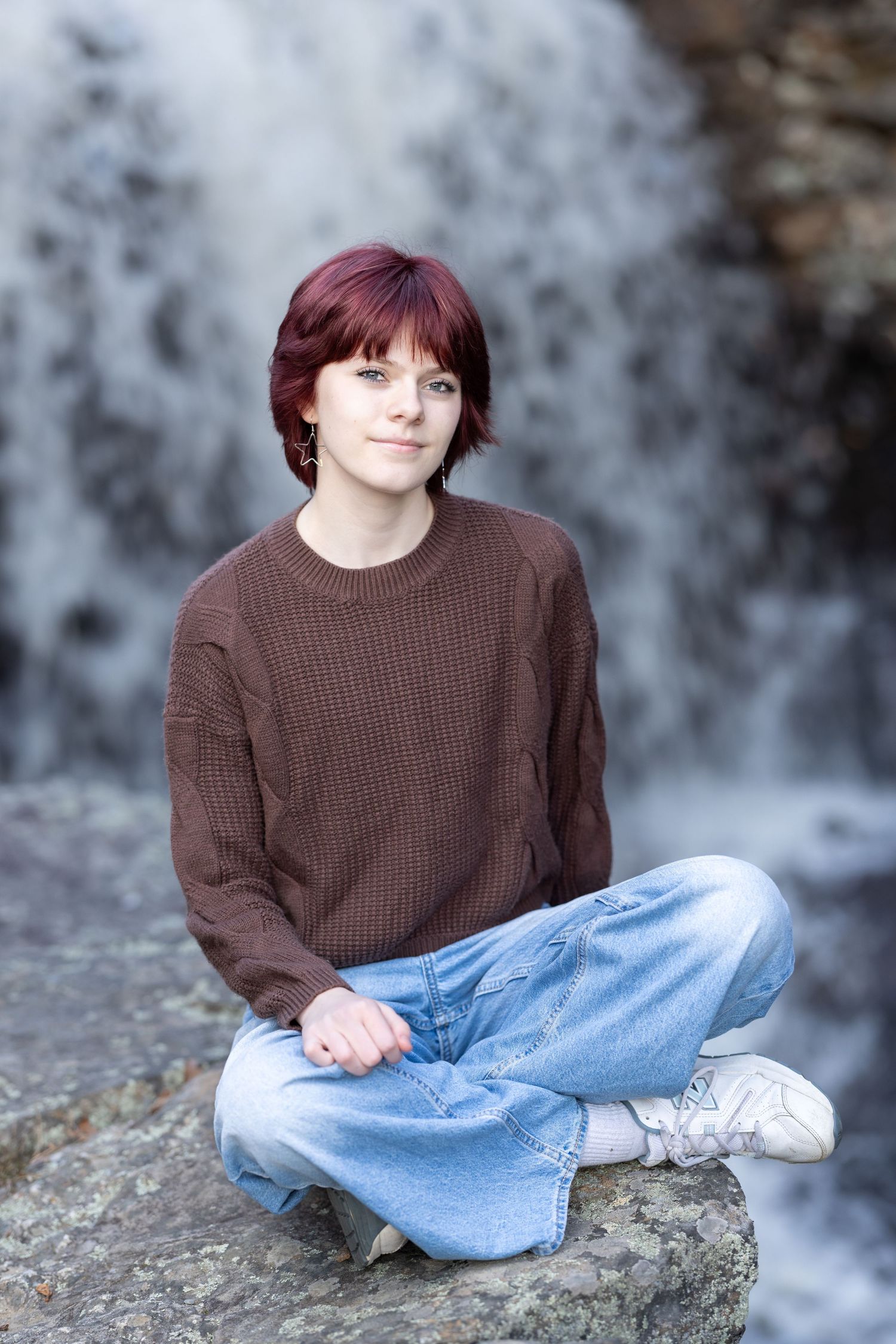 Person in brown sweater sitting on rock near waterfall wearing casual denim.