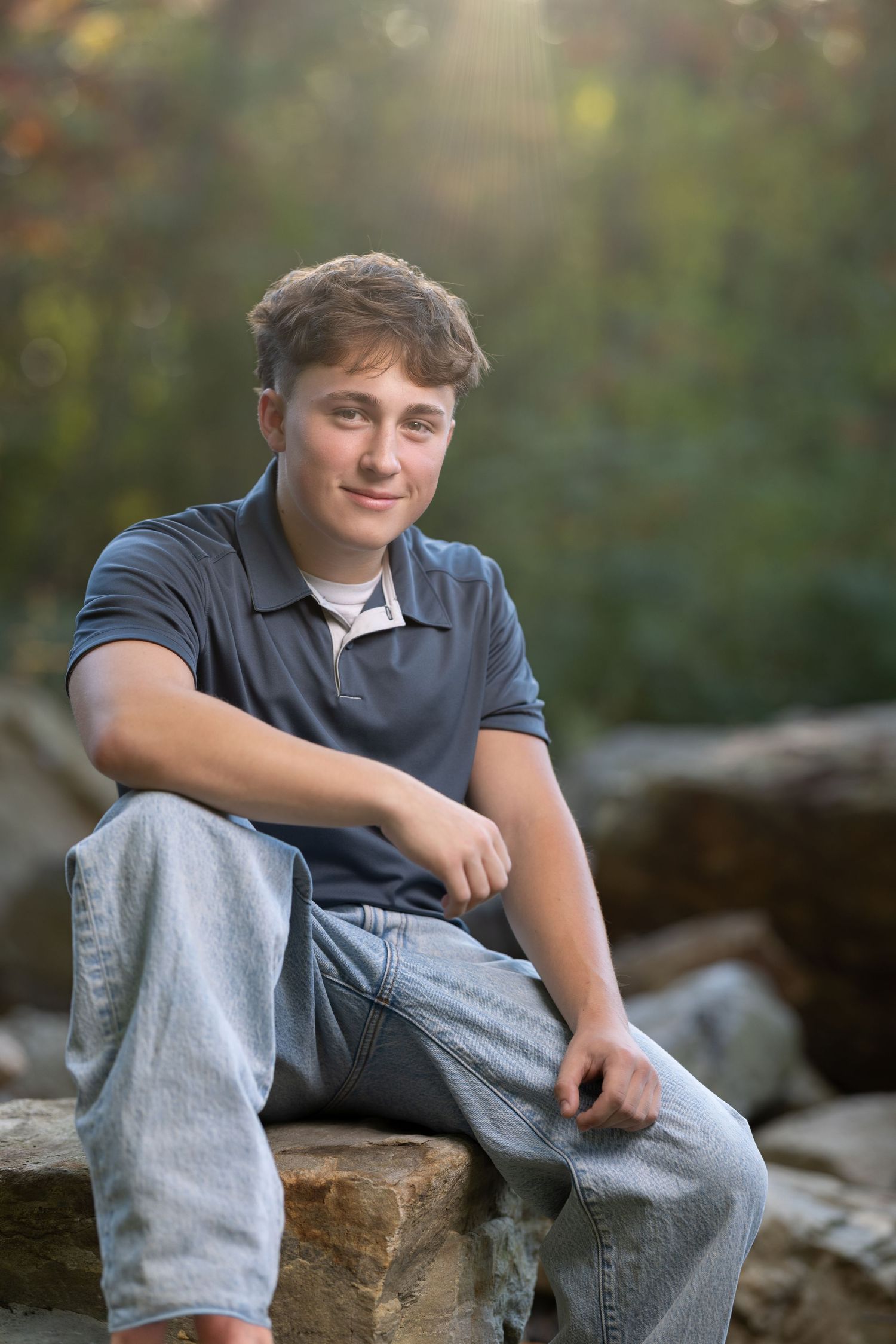 A young person sits casually on rocky terrain outdoors wearing gray pants and a dark shirt in natural lighting.