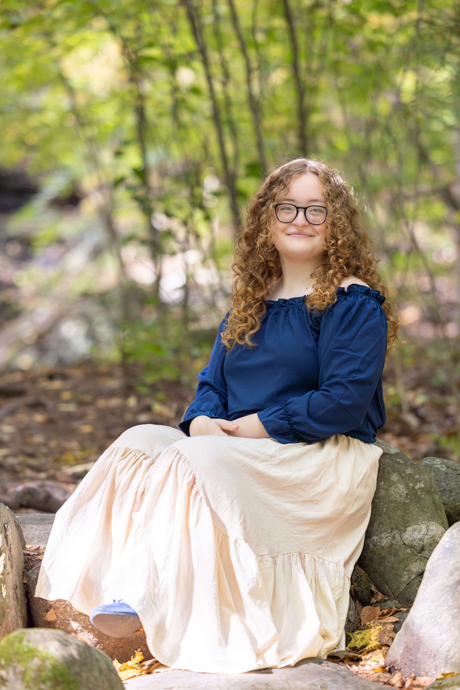 A person in a blue top and cream dress reclines on rocks in a wooded outdoor setting during an autumn photo session.