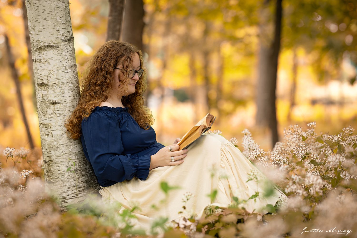A reader in a navy sweater sits against a birch tree surrounded by autumn foliage and wildflowers.