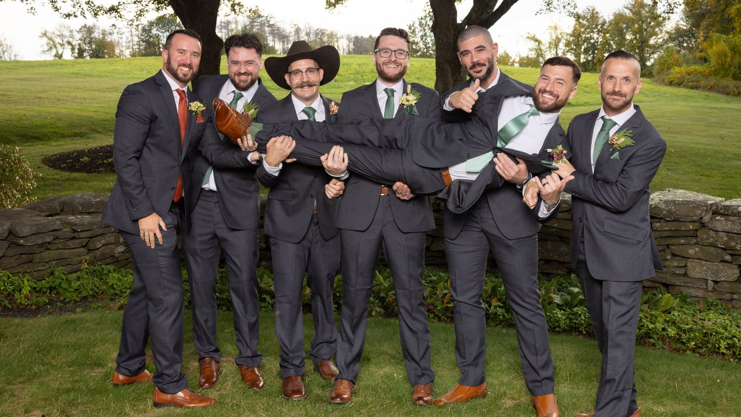Groomsmen in charcoal suits pose playfully in a row on outdoor stone steps at a wedding venue.