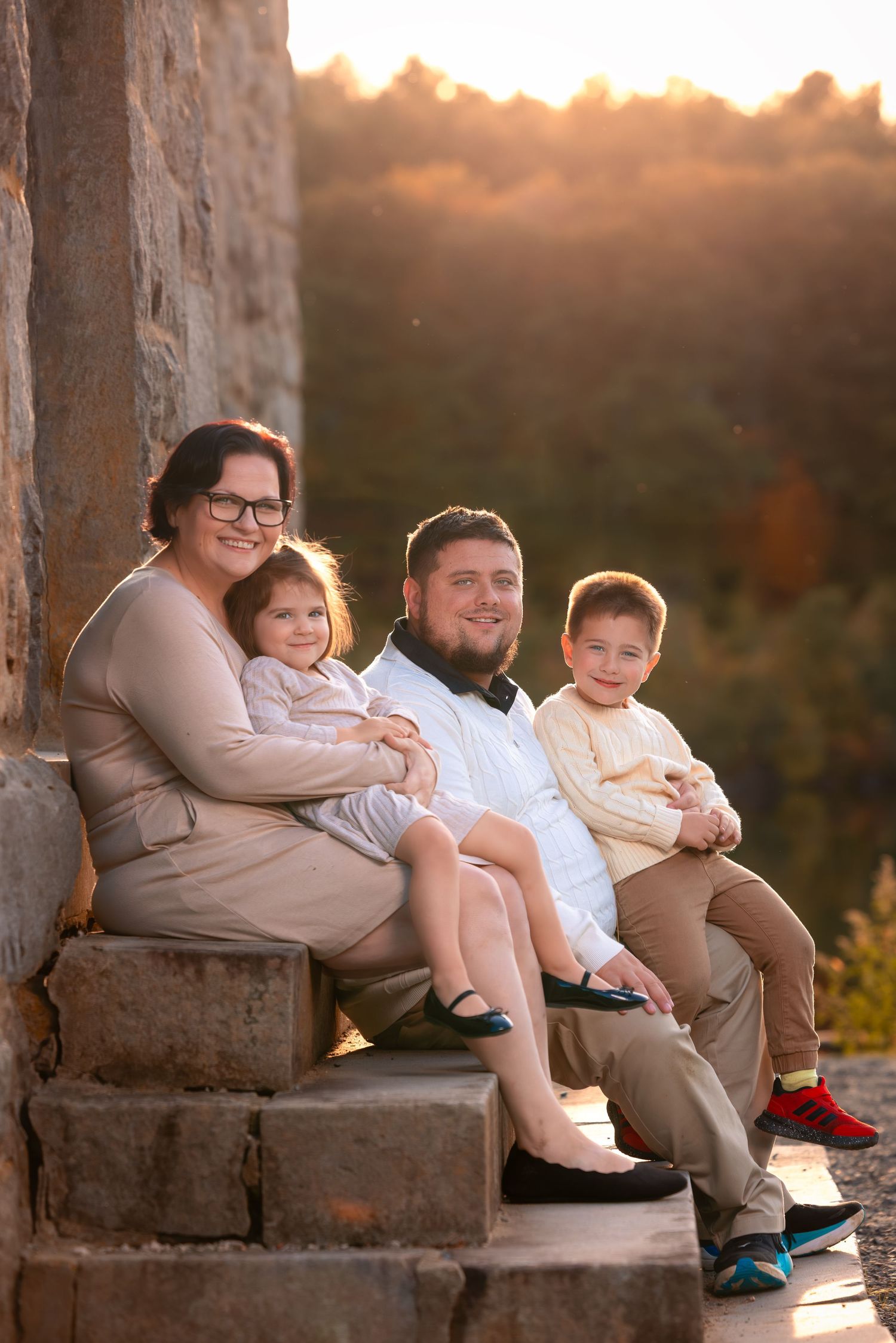 Family sits together on stone ledge during golden hour sunset in matching beige outfits.
