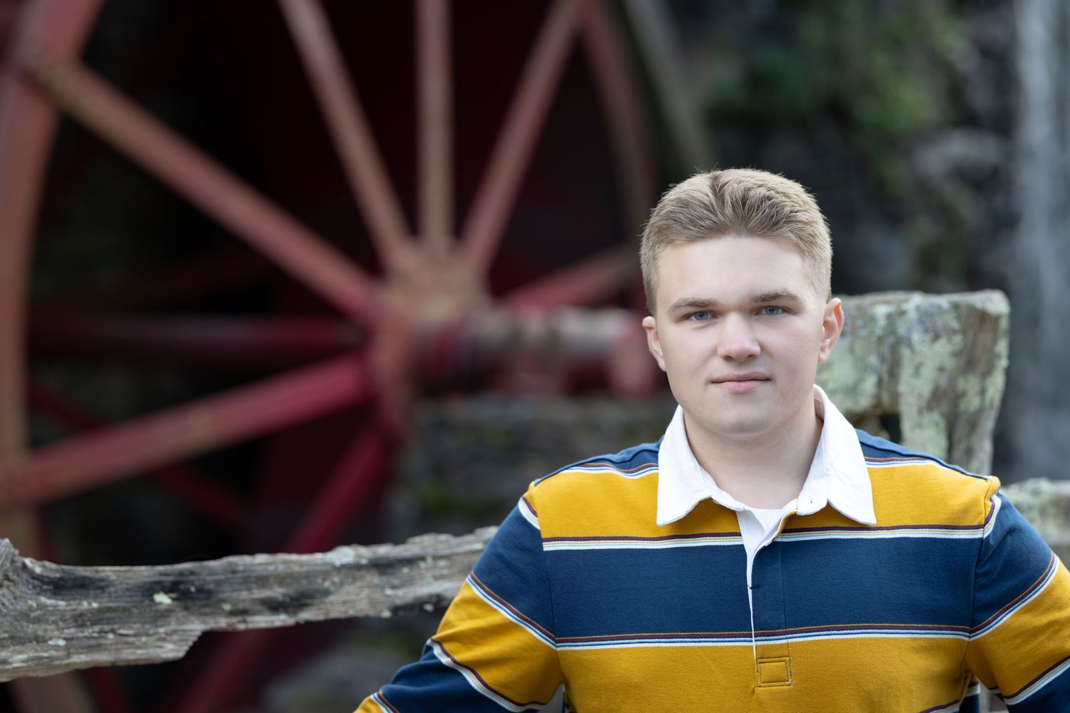 A person in a yellow and blue varsity jacket poses outdoors in front of a wooden structure.