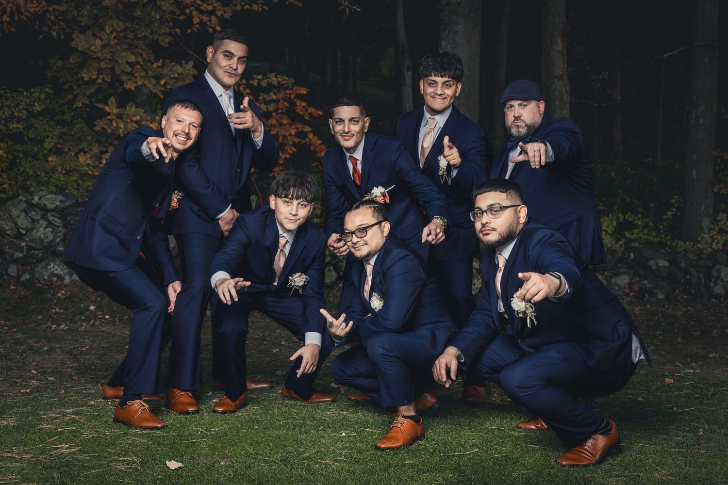 A wedding party in navy suits and brown shoes poses together in various casual positions on the grass.