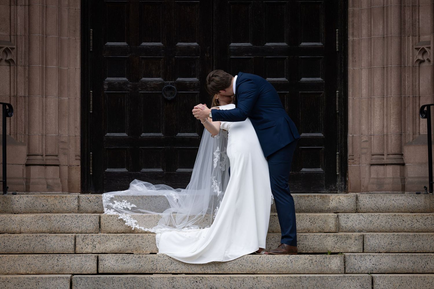 A bride and groom walk together along a grand stone building exterior with elegant columns and architecture.