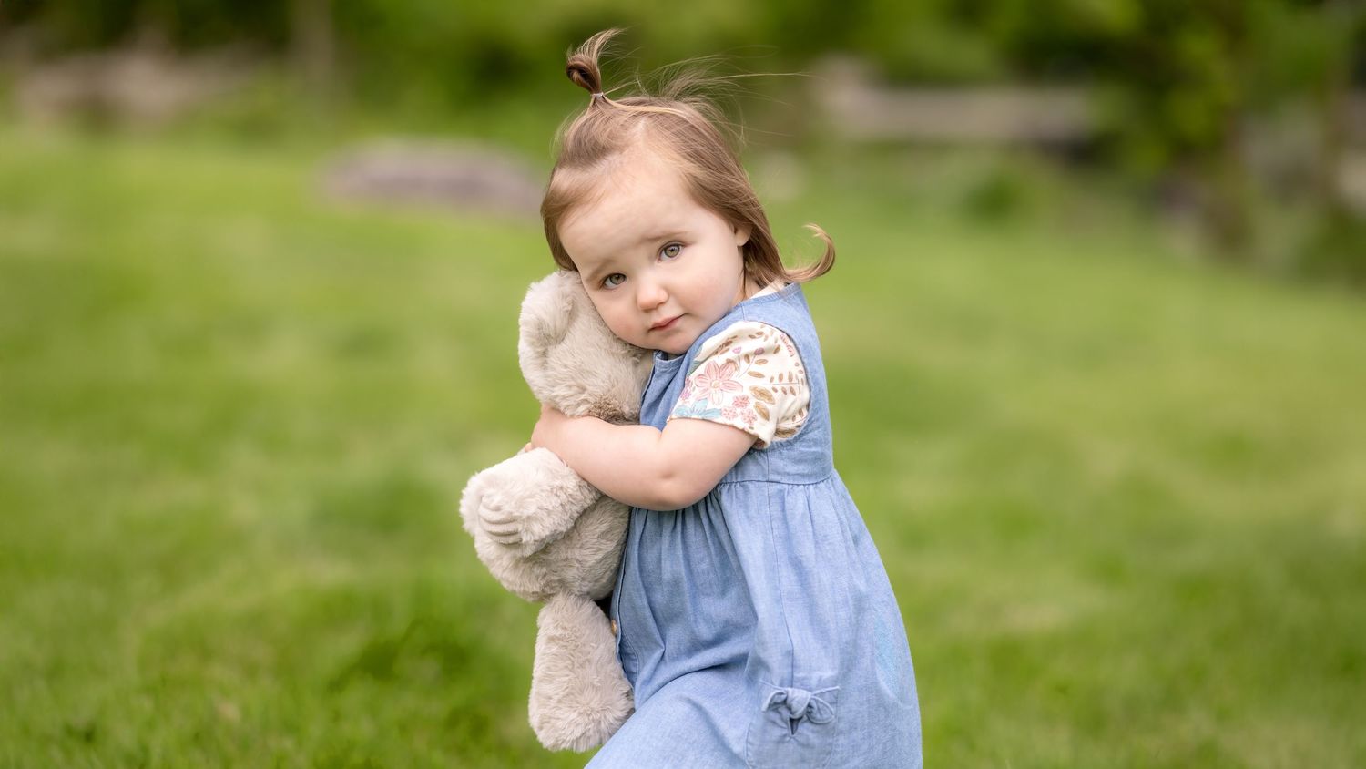 A toddler in a blue dress plays with a stuffed bunny toy in a grassy outdoor setting.