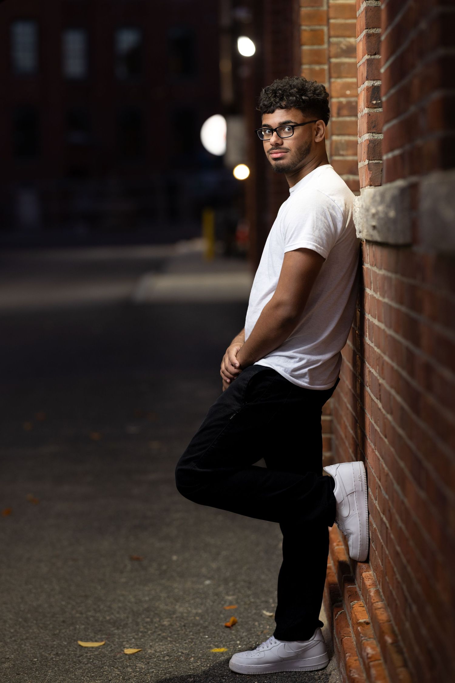 A person in a white t-shirt and black pants sits casually against a brick wall in an urban setting.