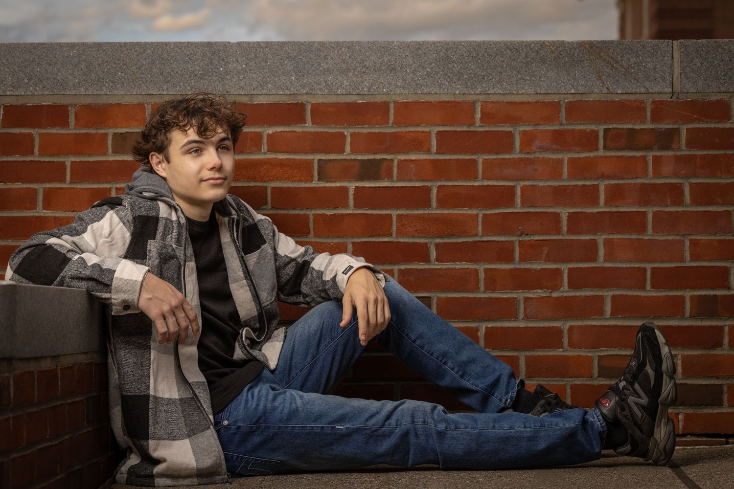 A person in a plaid jacket and jeans sits casually against a brick wall in an artistic photo series.