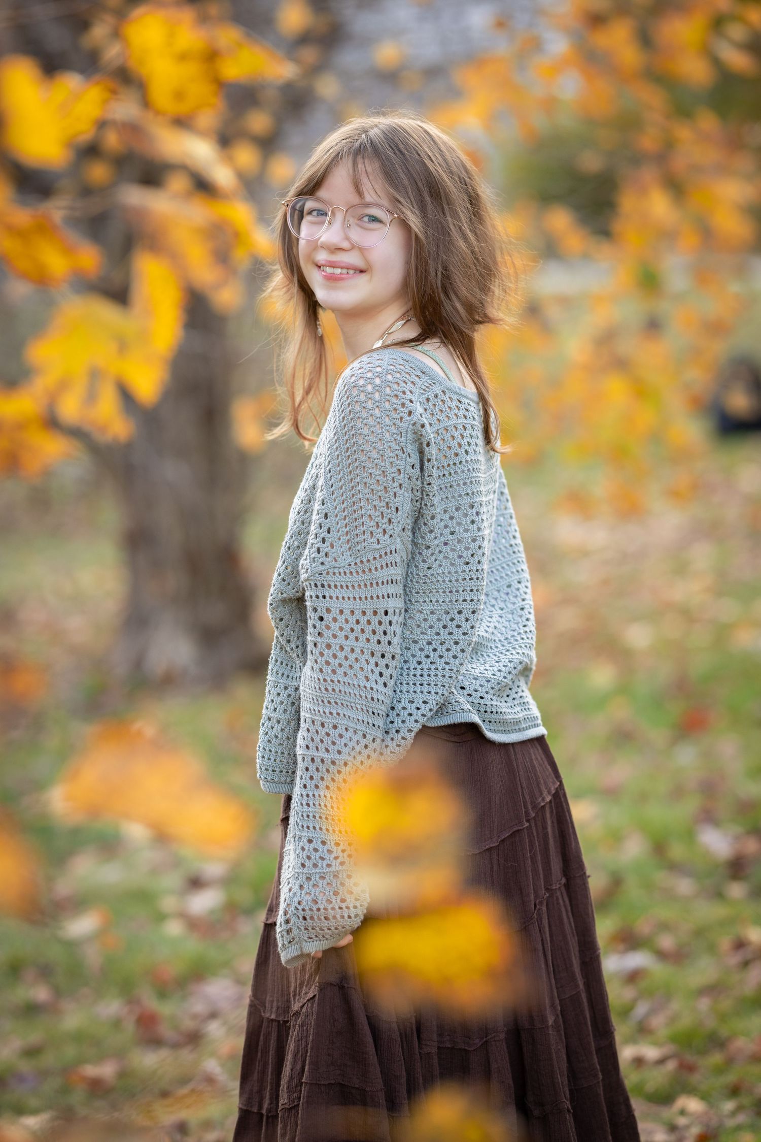 Child in a gray sweater and brown dress twirling among autumn leaves with golden foliage.