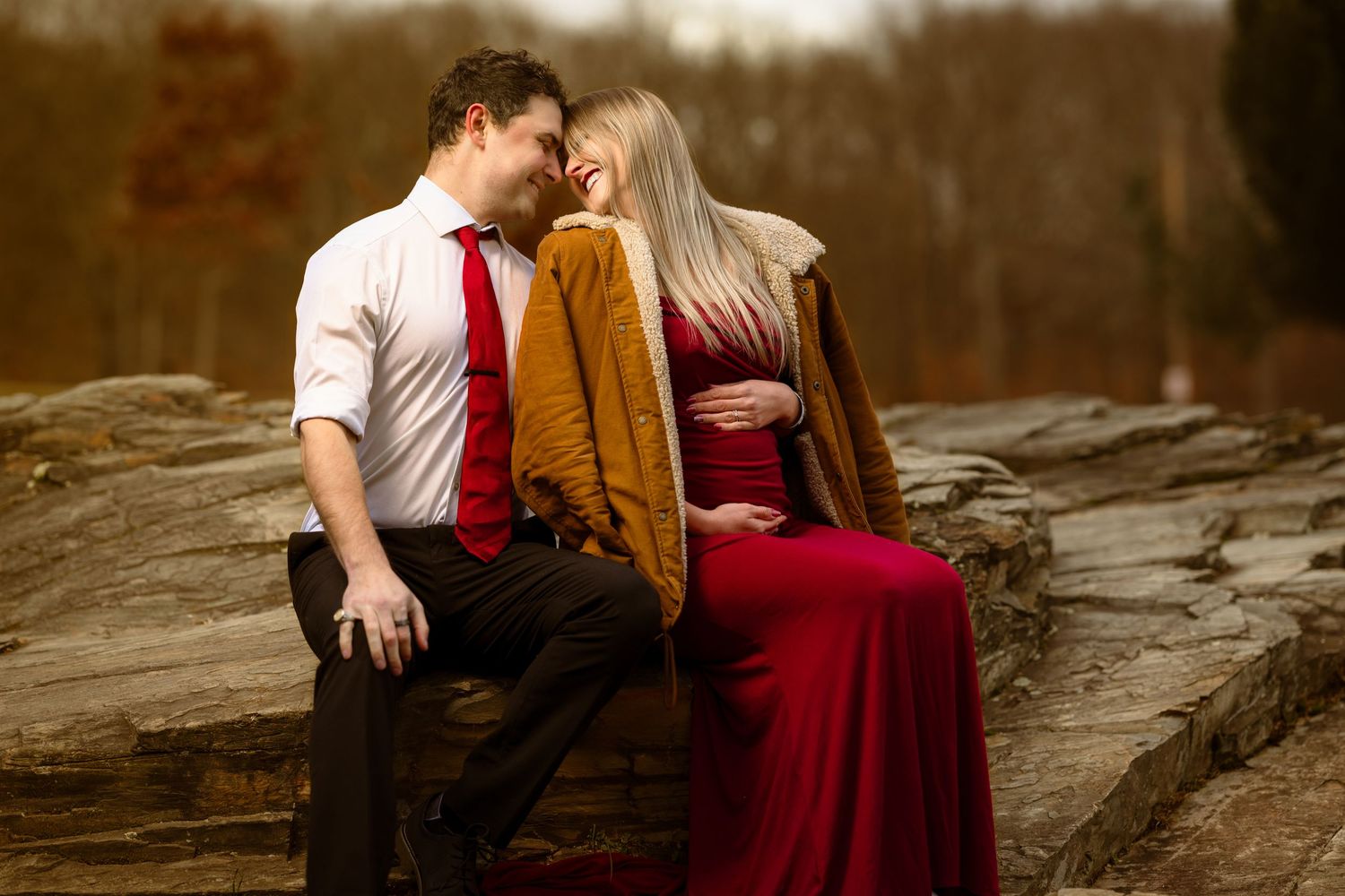 A couple in red and brown attire shares an intimate moment on a rocky surface during an autumn photoshoot.