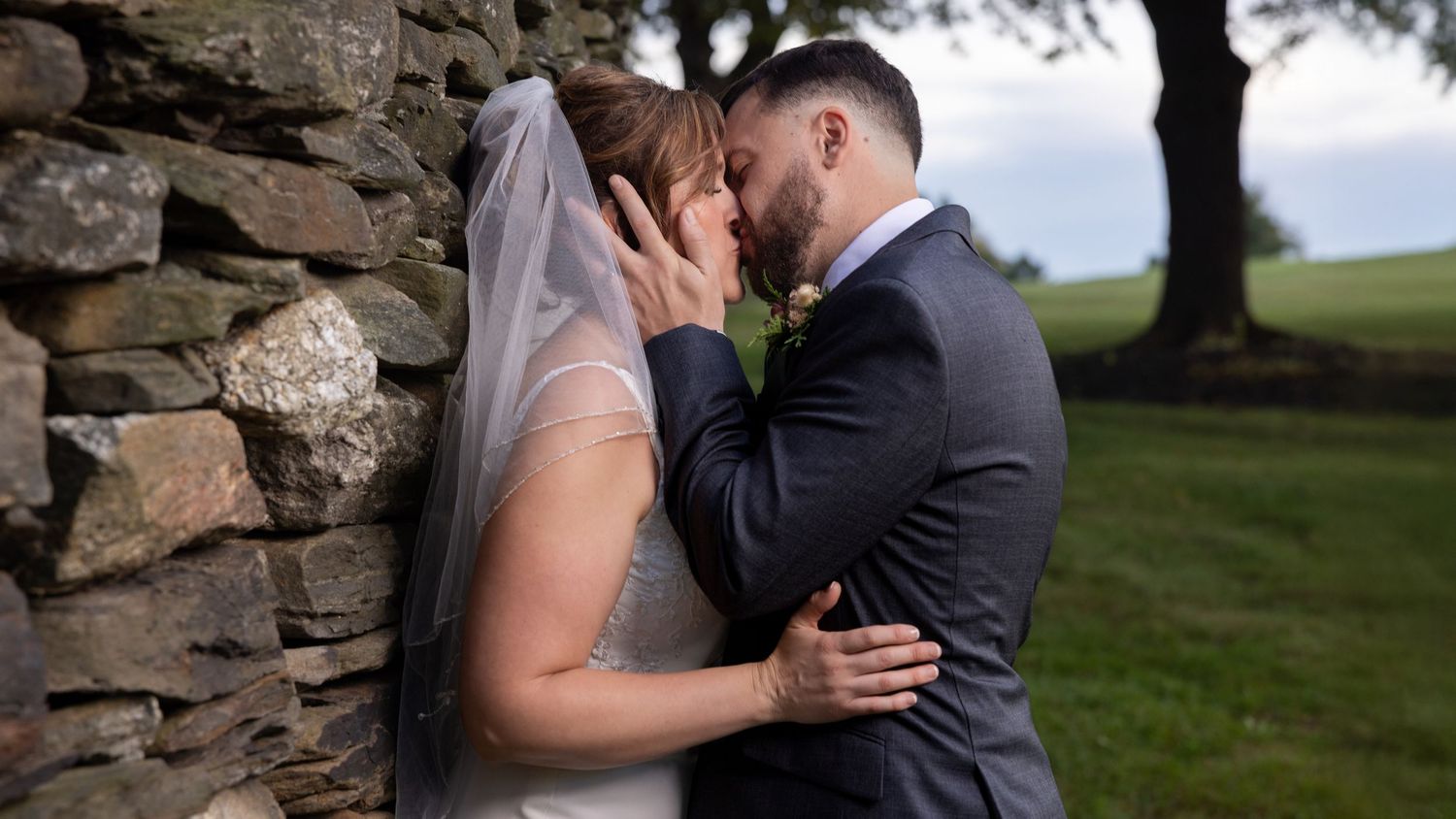 A romantic couple shares intimate moments by a stone wall during their wedding photoshoot in an outdoor setting.