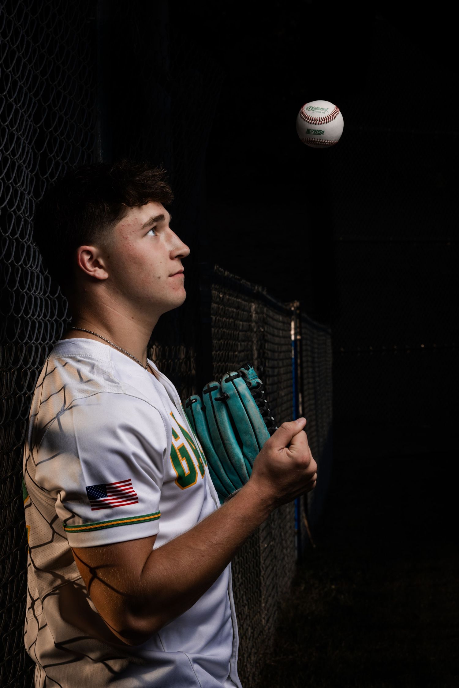 Baseball player in white uniform with teal glove watches baseball against dark background with dramatic lighting.