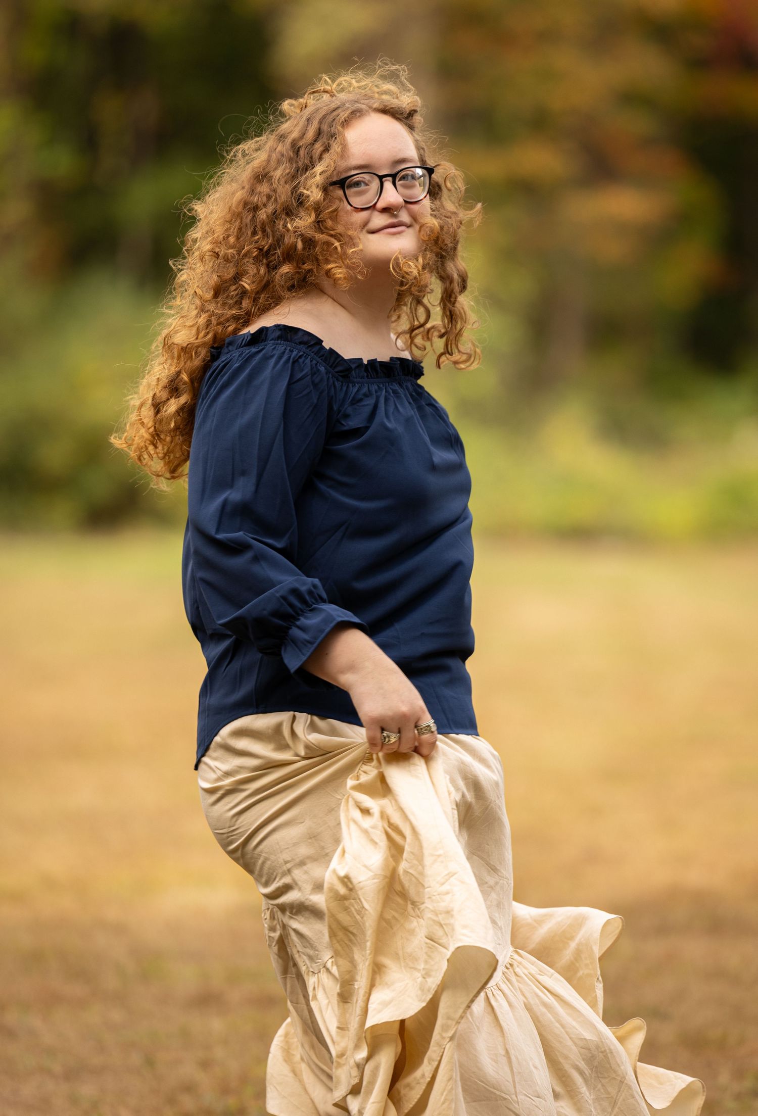 A person in a navy blue top and beige skirt twirls joyfully in an outdoor autumn setting.
