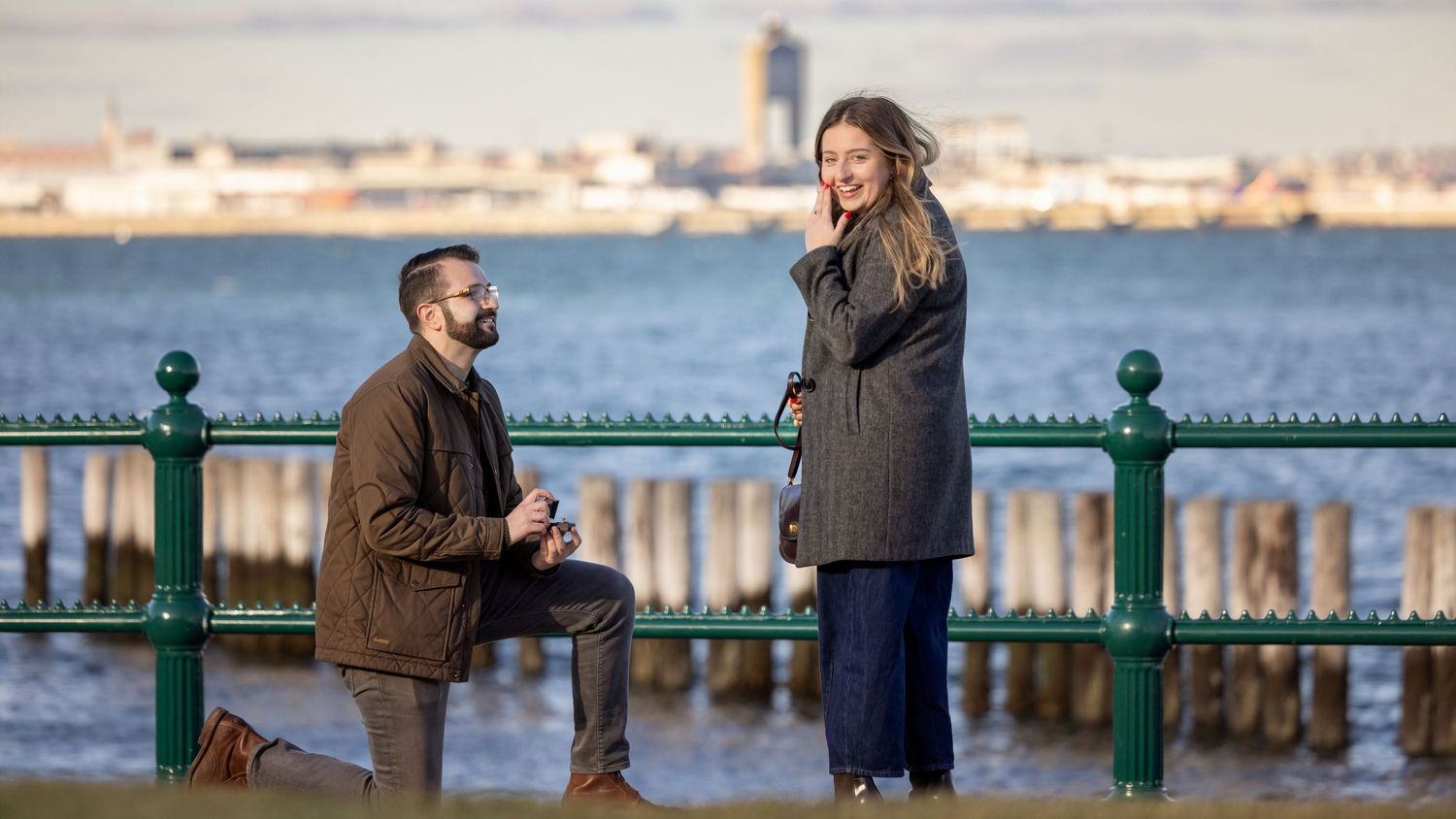 Couple walking along a waterfront pier with green railings and city skyline in the background.
