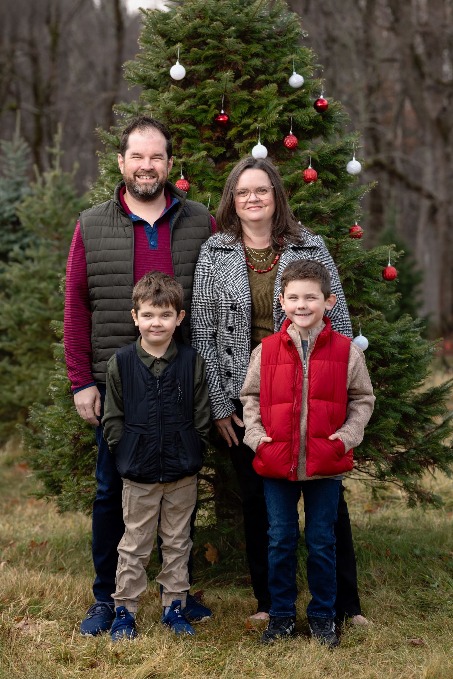 Family poses together in front of decorated Christmas tree outdoors in wooded area.