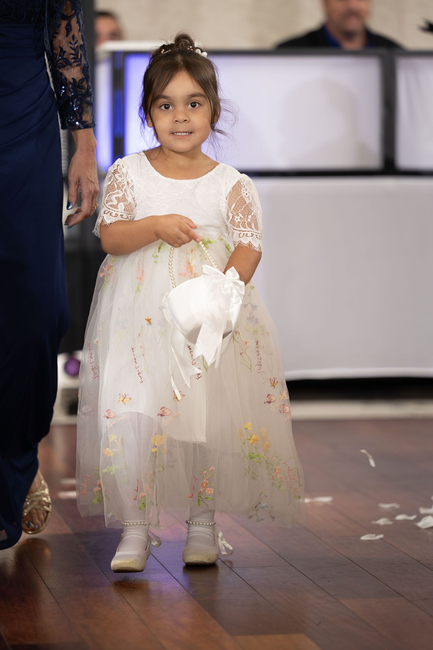 A small child in a white lace dress walks across a wooden floor during what appears to be a formal event.