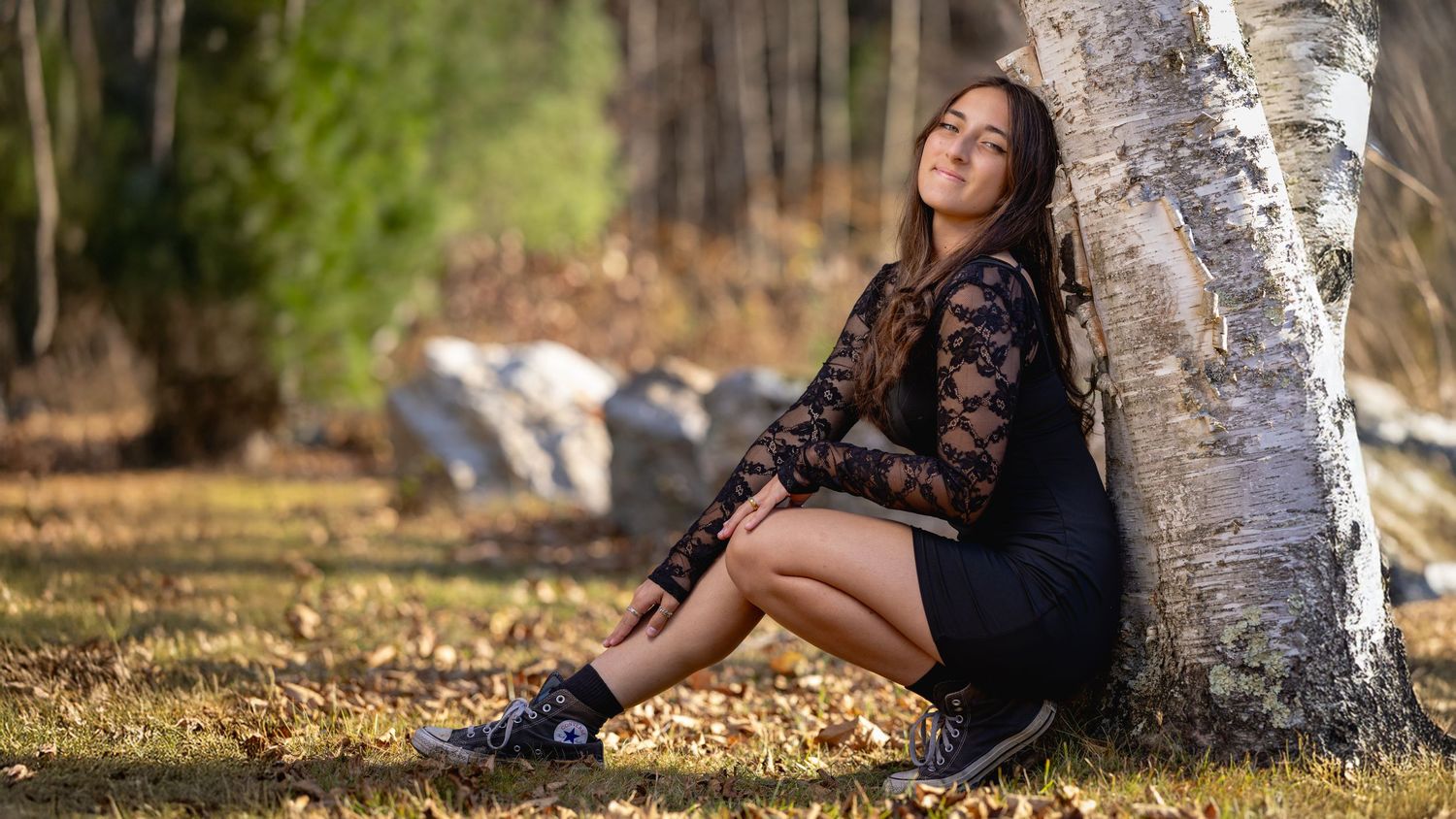 A person in a black dress sits against a tree trunk in a fall setting with golden leaves scattered on the ground.