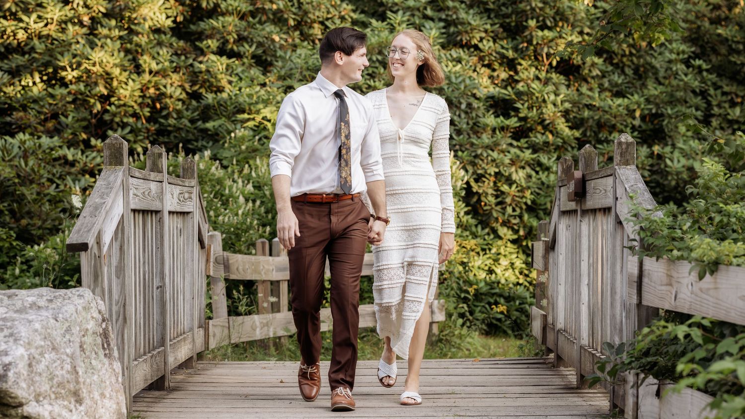 A couple walks hand in hand along a rustic wooden bridge surrounded by lush greenery on a sunny day.