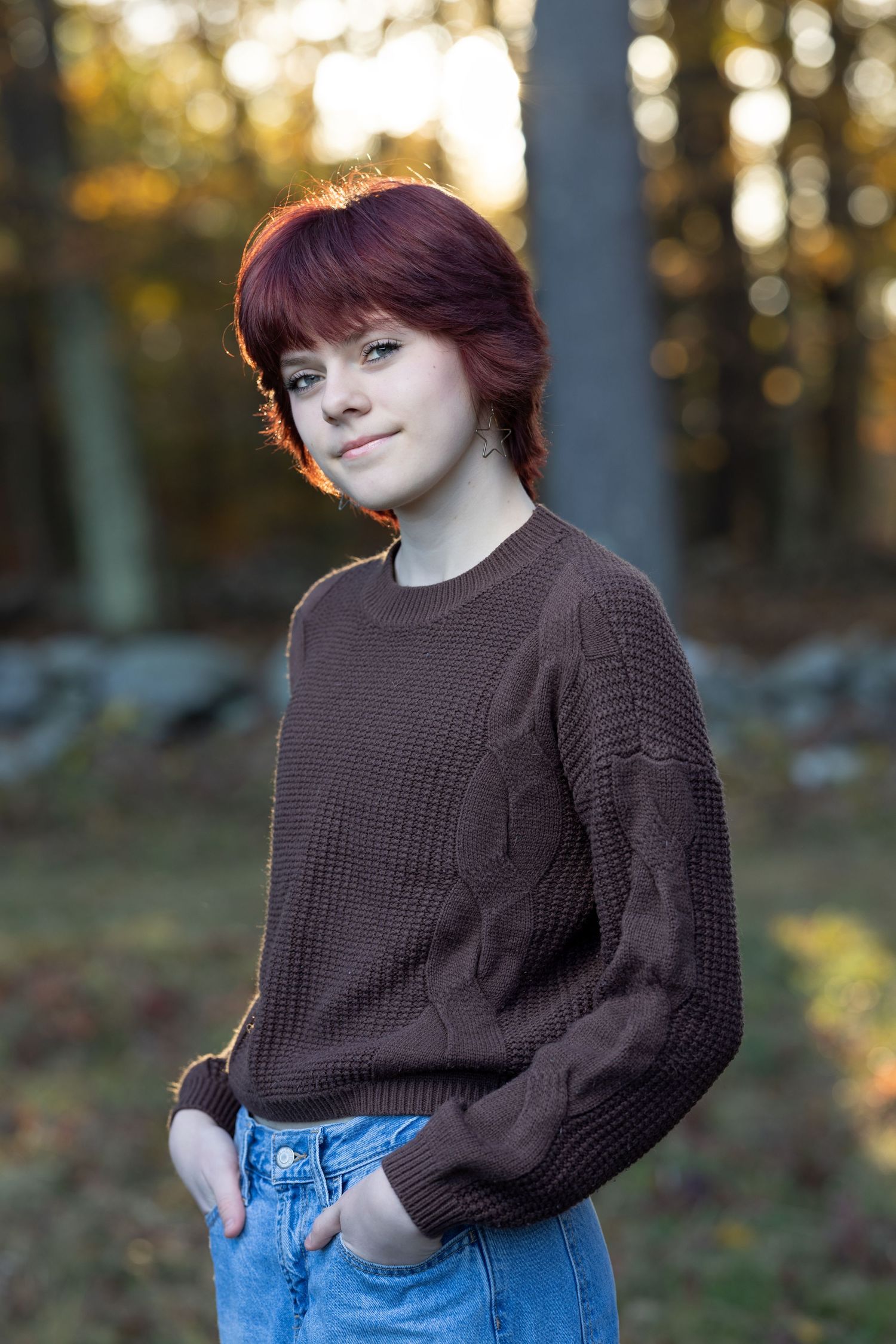 A person wearing a brown knit sweater and blue jeans poses outdoors in autumn lighting.