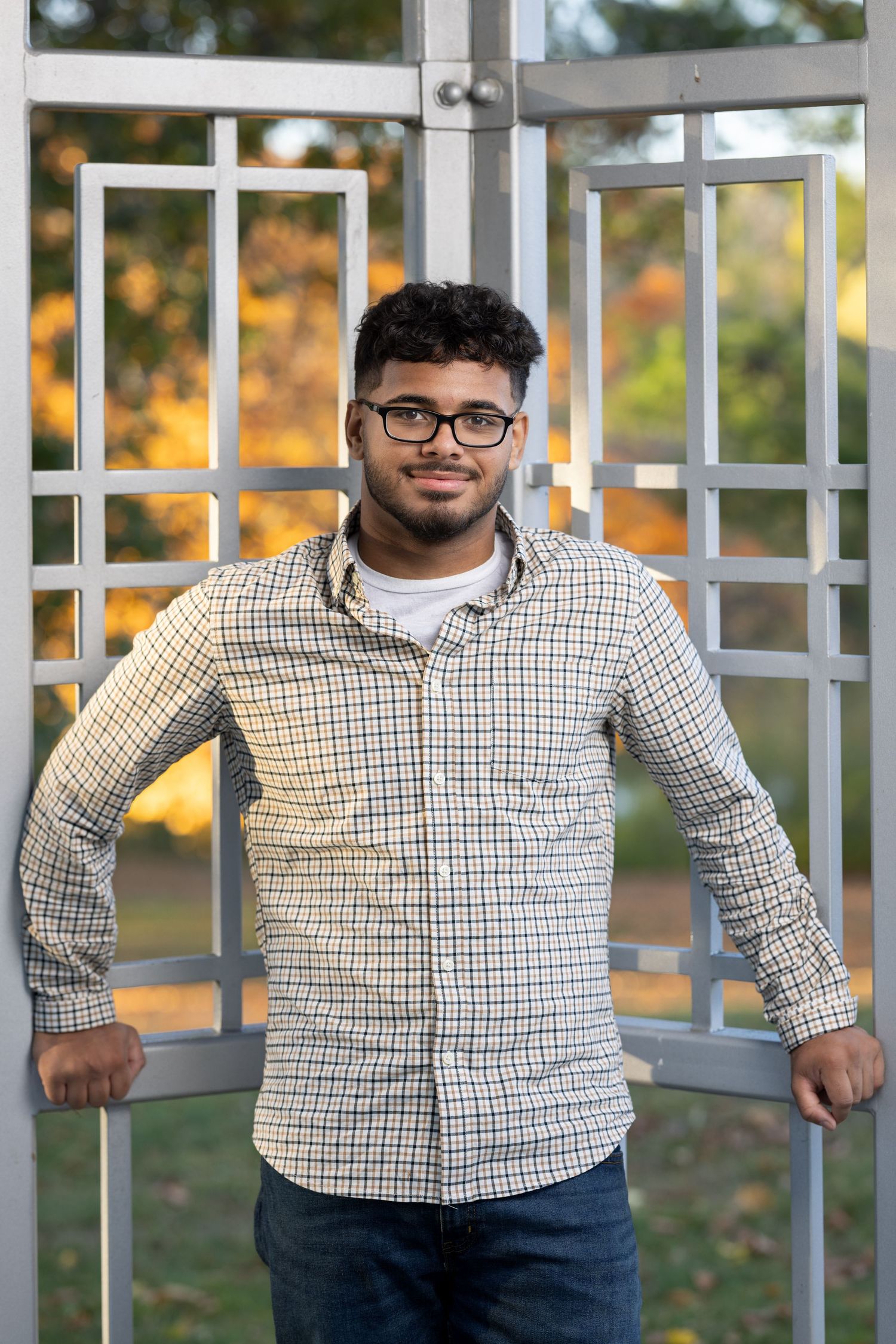 Person in a checkered button-down shirt and jeans poses against a metal railing with autumn foliage in background.