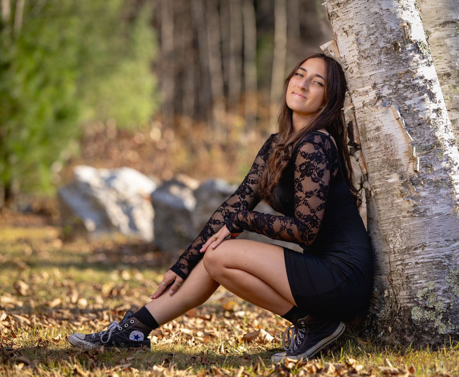 A person in black lace dress poses against a birch tree trunk surrounded by fallen autumn leaves in golden light.