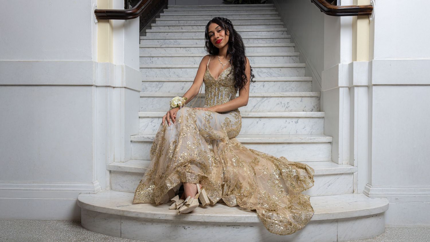 A figure in a flowing champagne-colored gown poses elegantly on white stairs in a classic architectural setting.