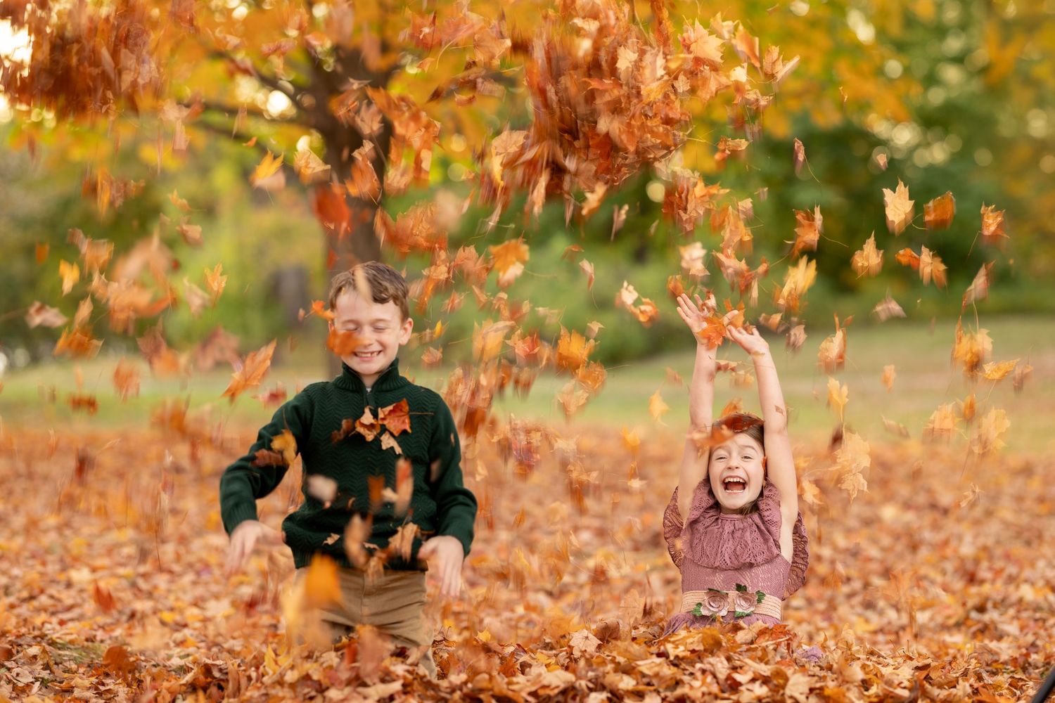 Children play and throw autumn leaves in the air during a fall photoshoot in a park setting.