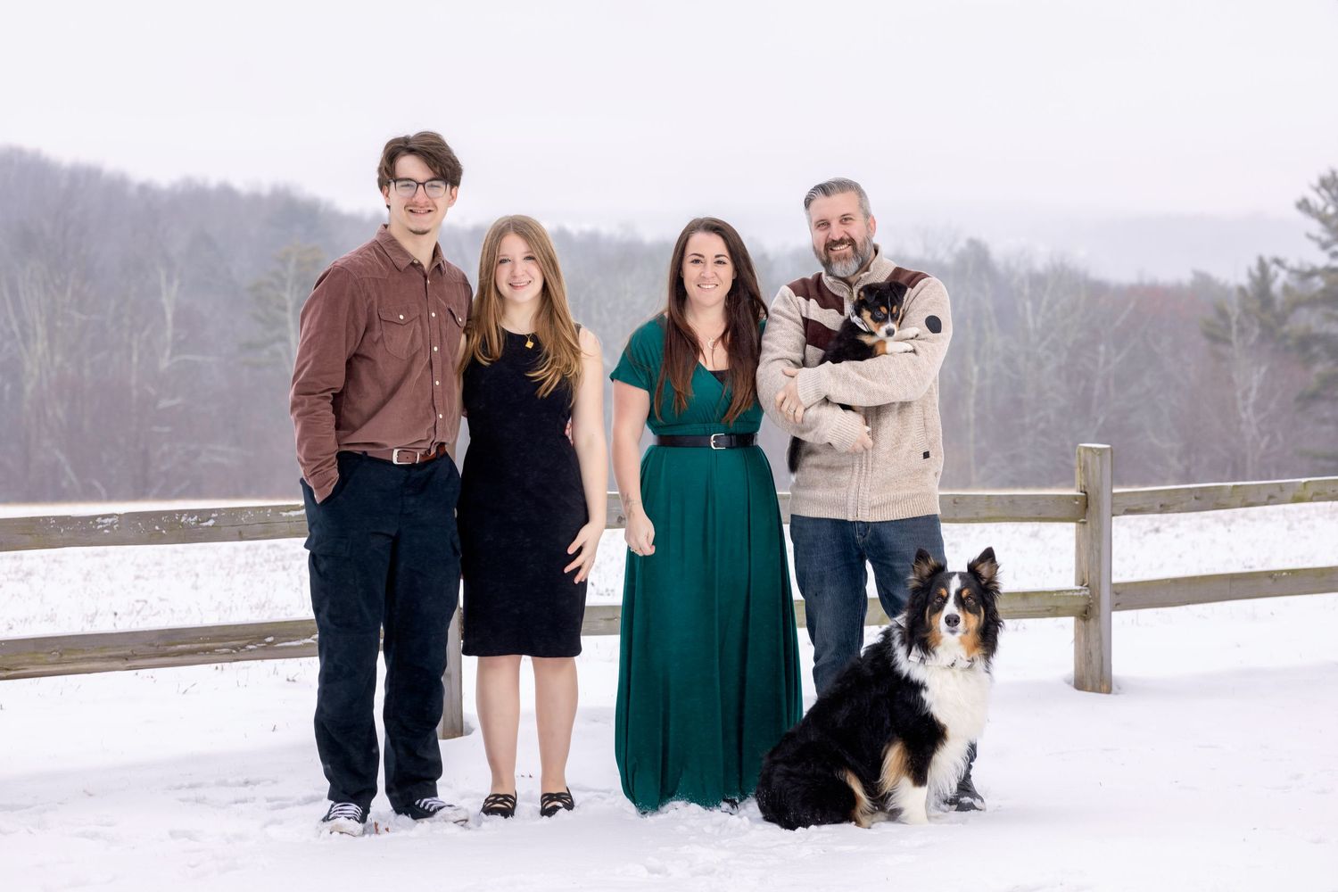 Family wearing coordinated outfits poses together in winter setting along wooden fence during outdoor photo session.