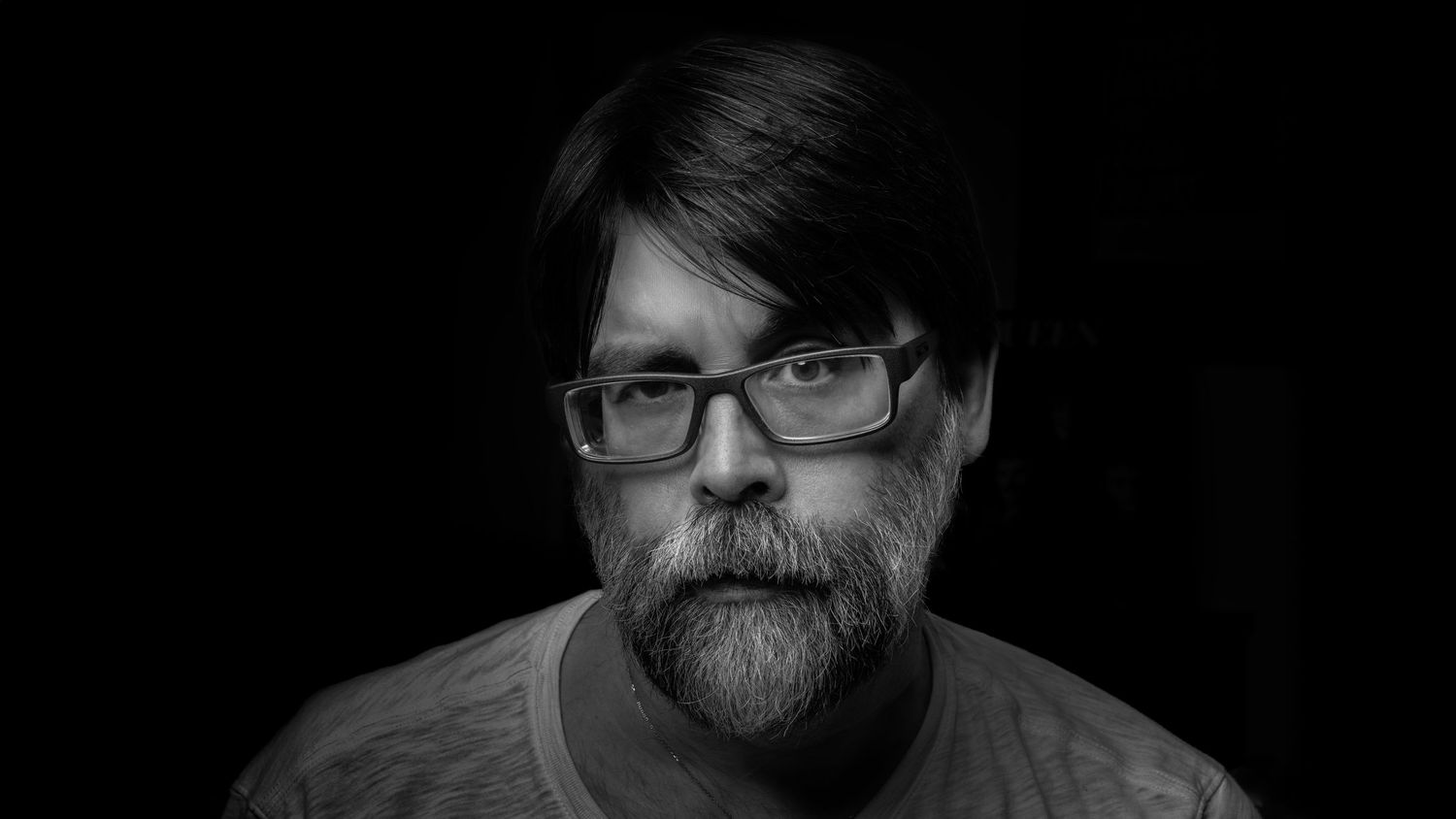 Black and white portrait of person with glasses, beard and side-swept hair against dark background.