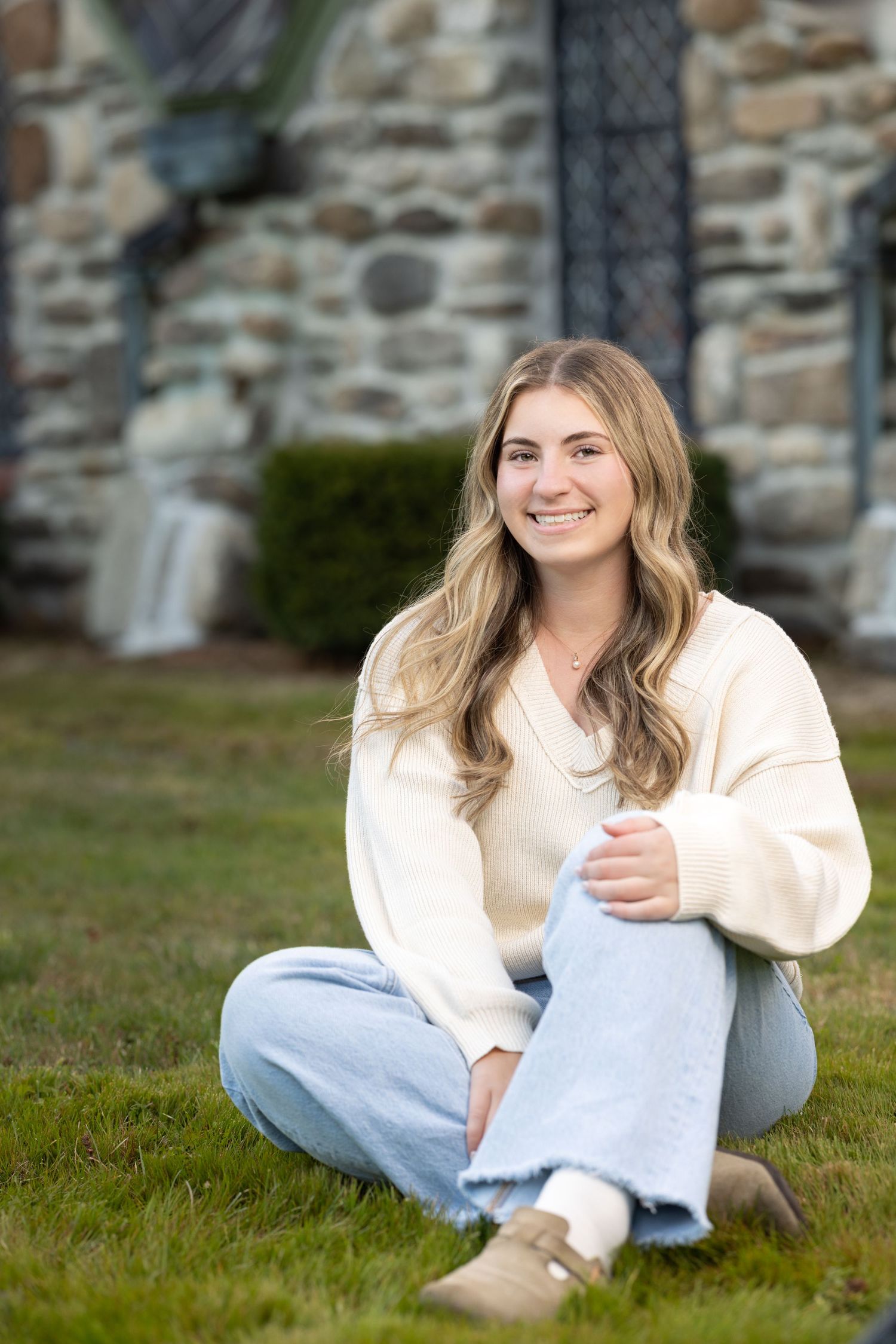 A person in a cream sweater and light jeans sits casually on grass near a stone wall.