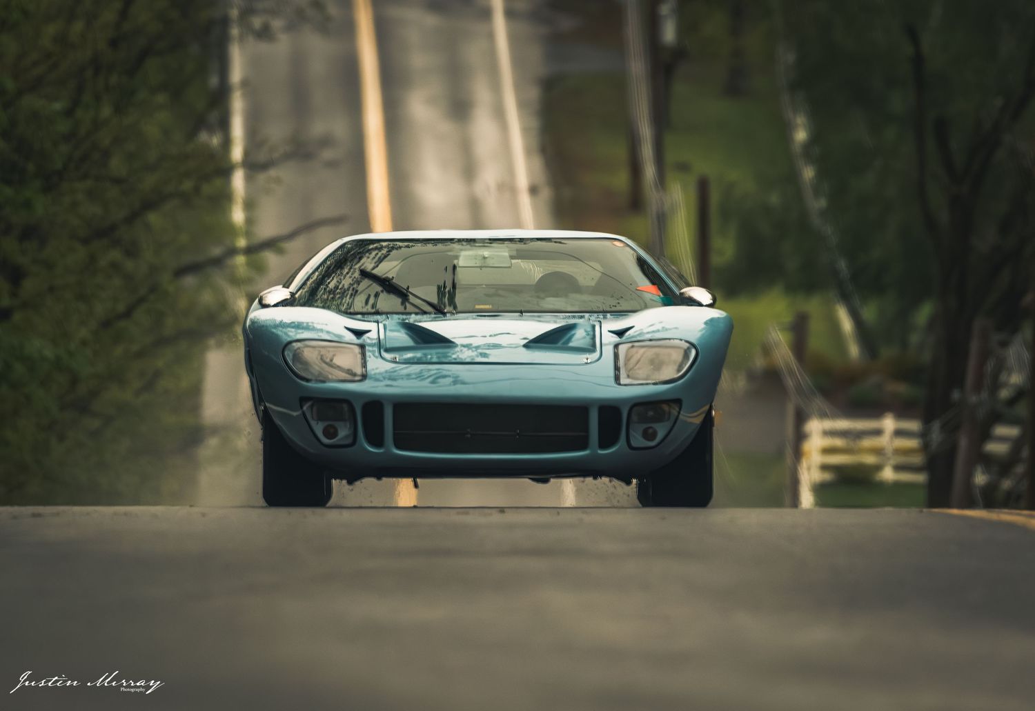 A light blue Ford GT supercar drives on a tree-lined road in dramatic lighting conditions.