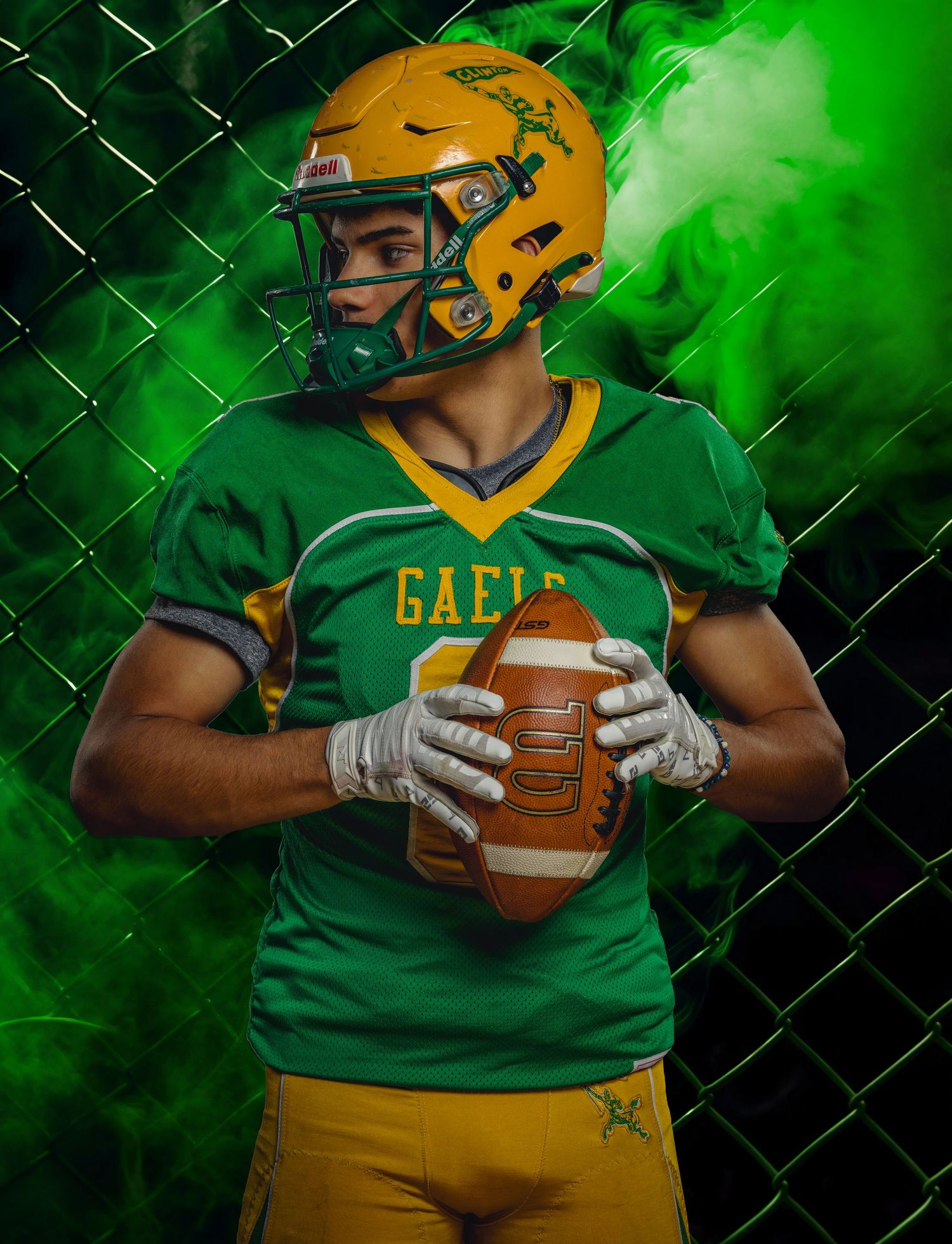 Football player in green and gold uniform holds ball against chain-link fence with dramatic green lighting effects.