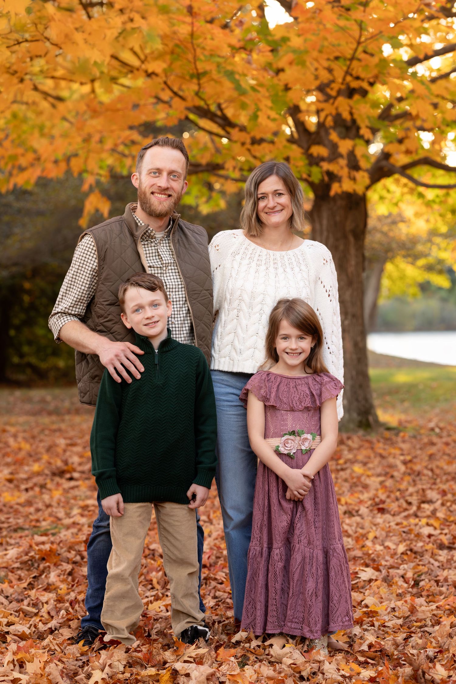 A family of four poses together on fallen autumn leaves with golden trees in the background during sunset.